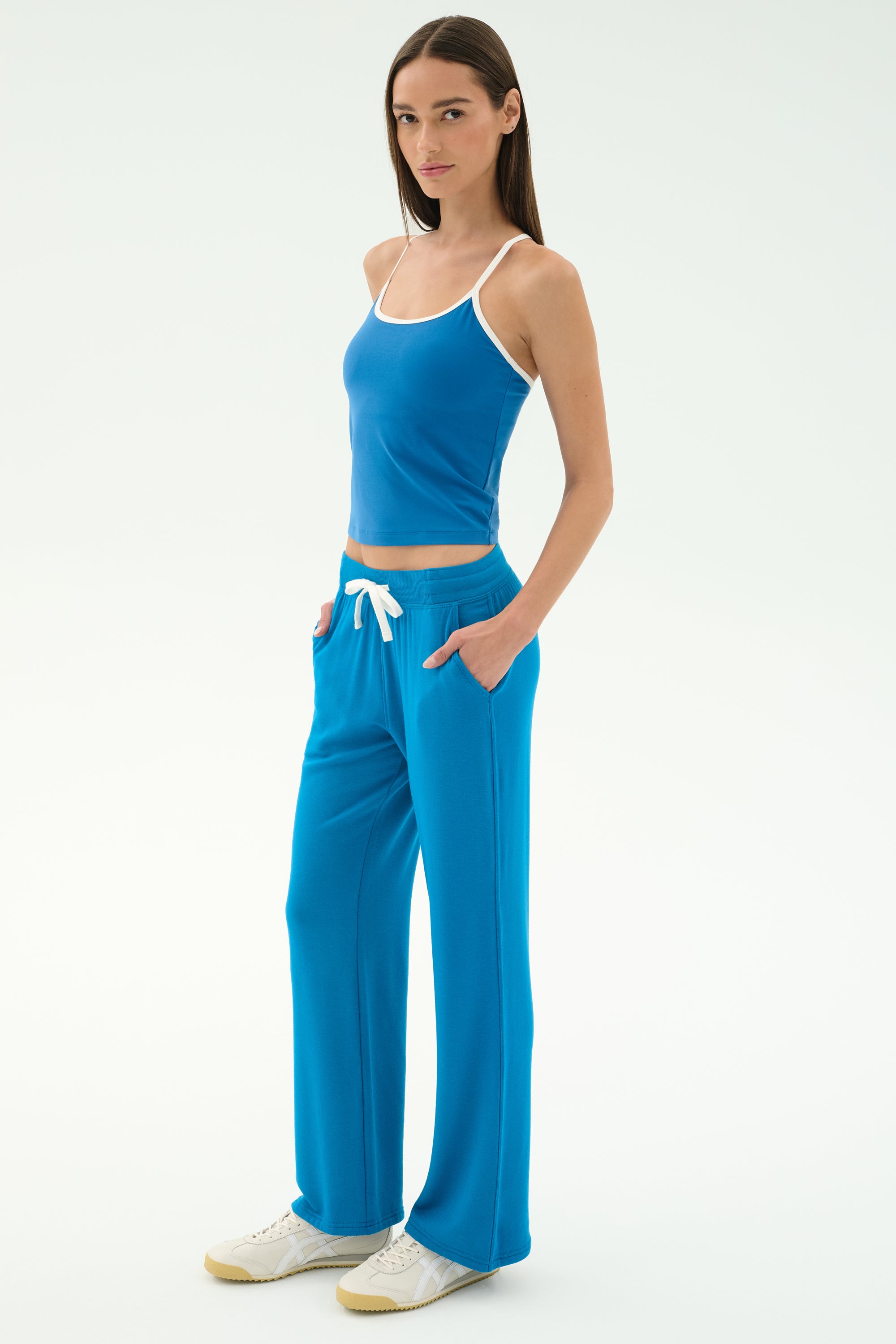 A woman stands against a plain background in the SPLITS59 Airweight Tank—Stone Blue/White—with matching drawstring pants and white sneakers, hands in pockets, ready for Pilates or hot yoga.