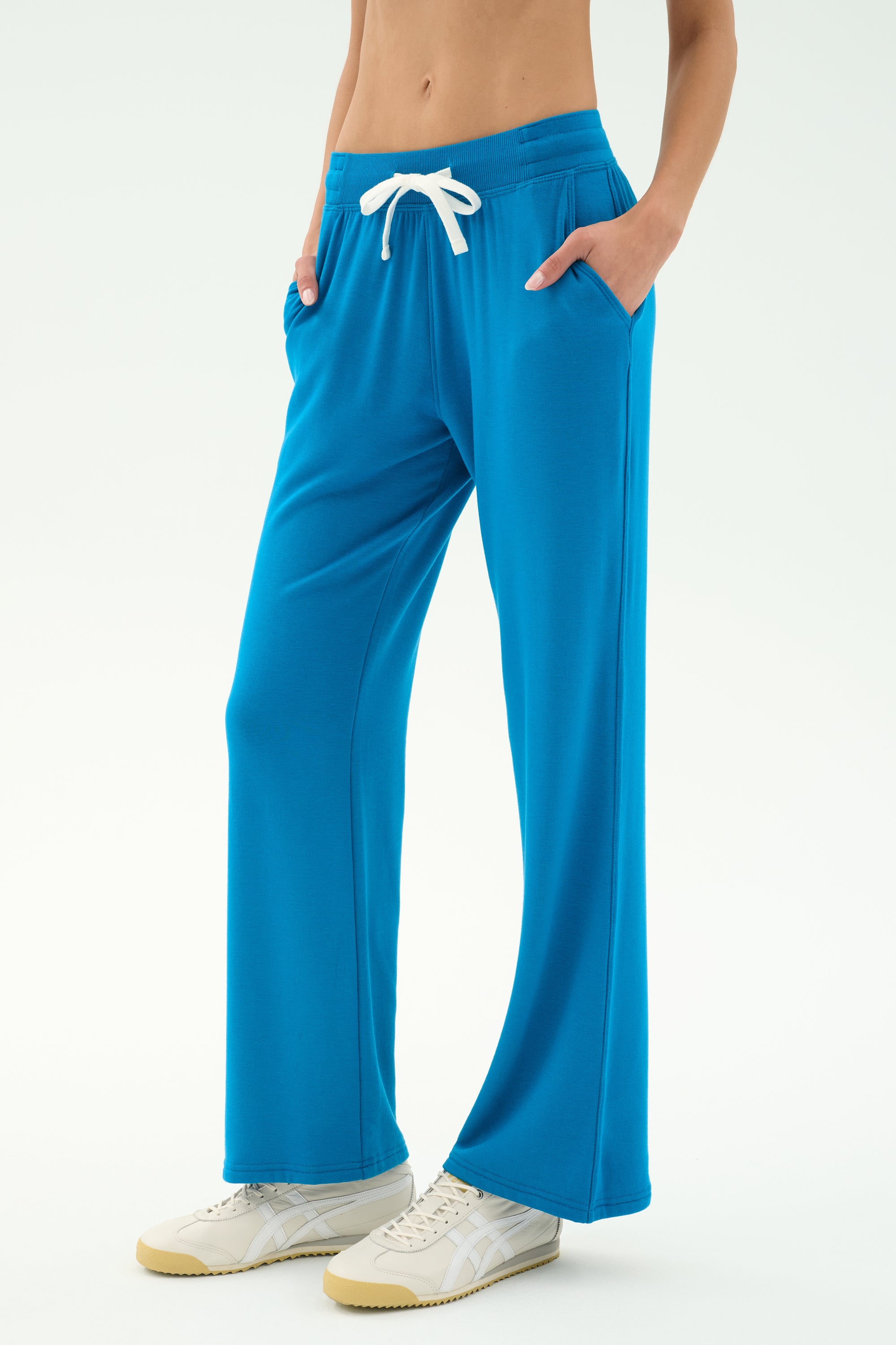 A person wears SPLITS59's Raven Fleece Full Length Sweatpant in Stone Blue—luxury Los Angeles-made ultra-luxe fleece—with white sneakers, hands in pockets against a plain background.