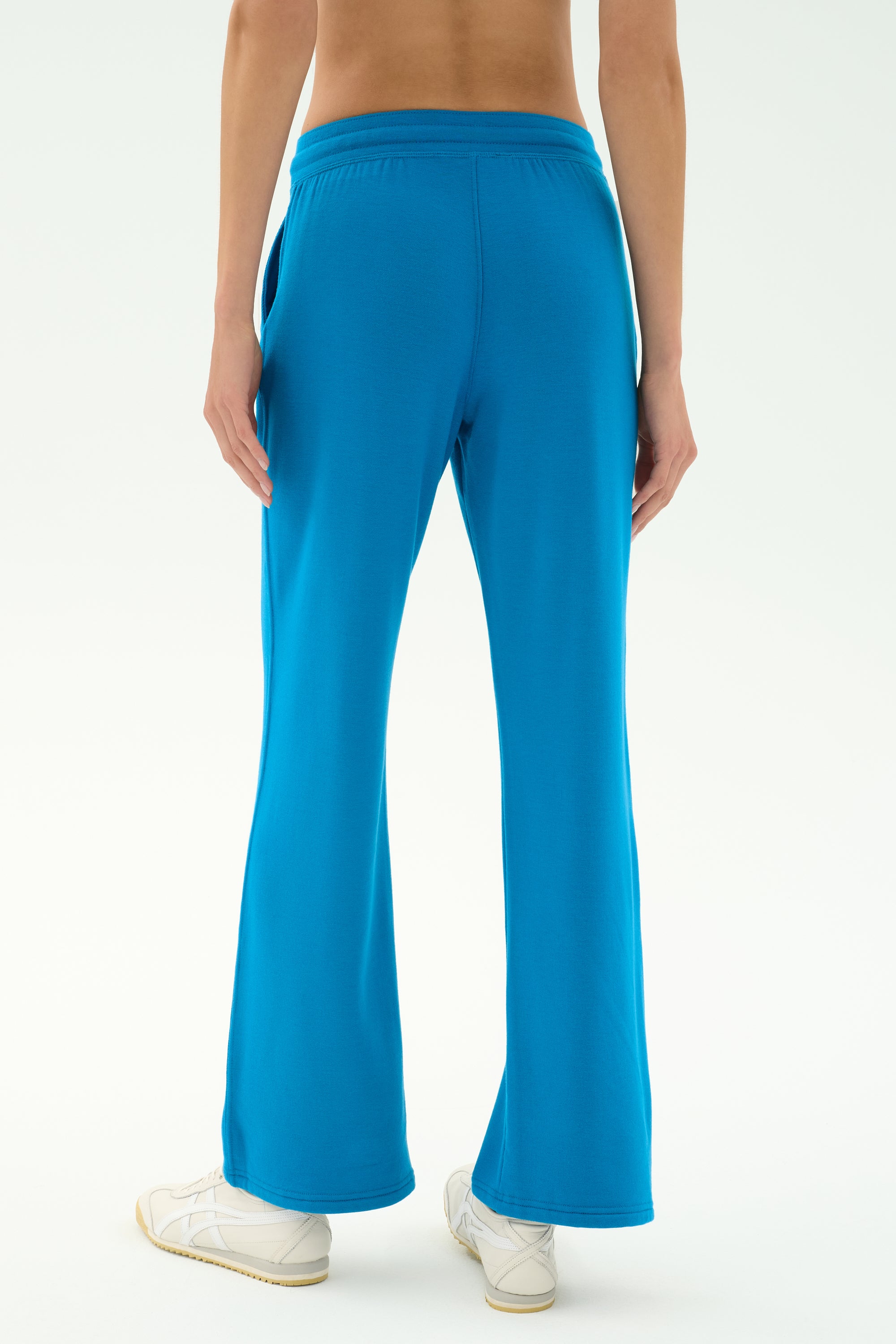 A person wearing SPLITS59 Raven Fleece Full Length Sweatpant in Stone Blue and white sneakers is shown from the back against a plain light background.