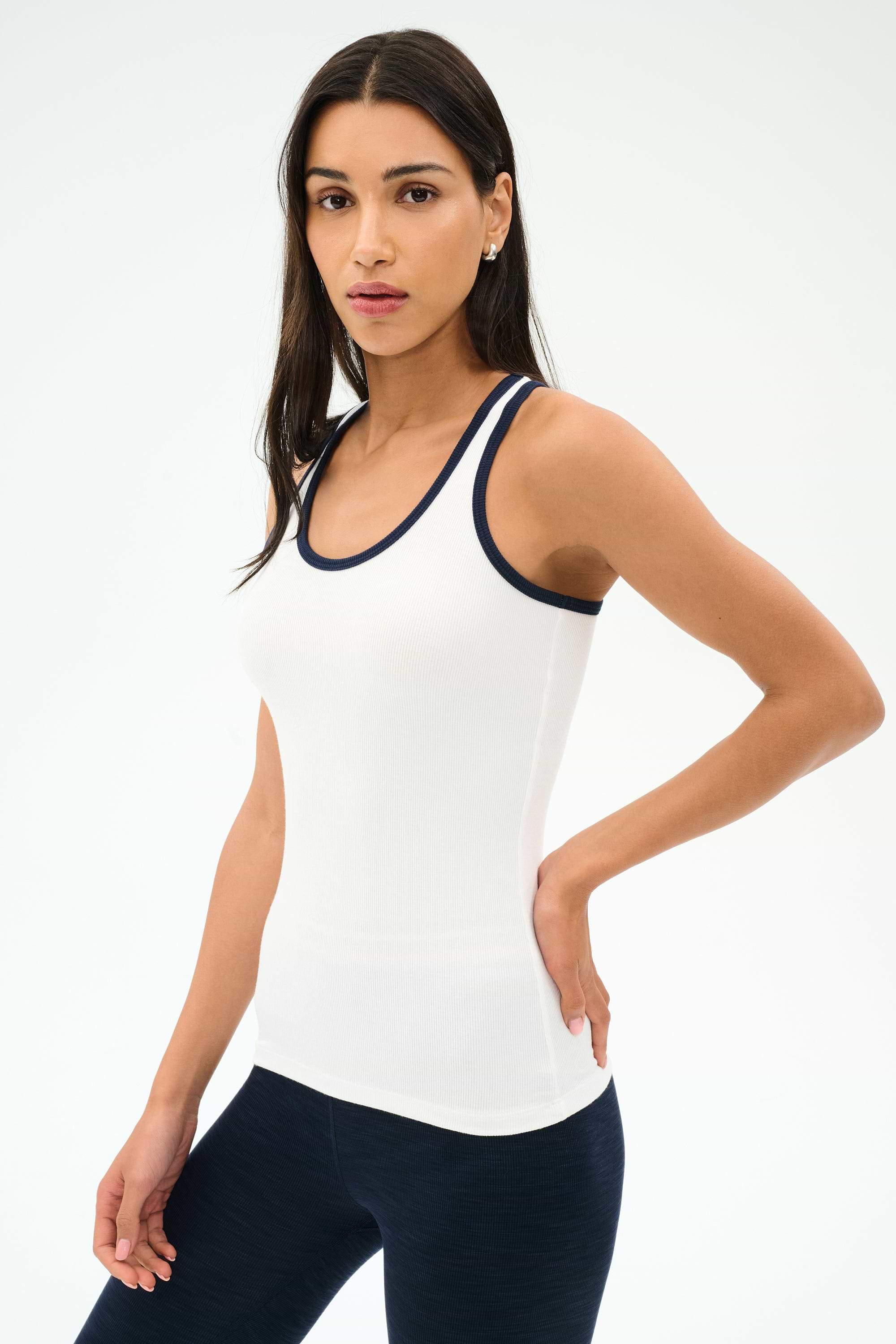 A woman stands against a plain background, wearing the SPLITS59 Ashby Rib Tank in White/Indigo and navy leggings, looking at the camera with a neutral expression—ideal for yoga or gym workouts.