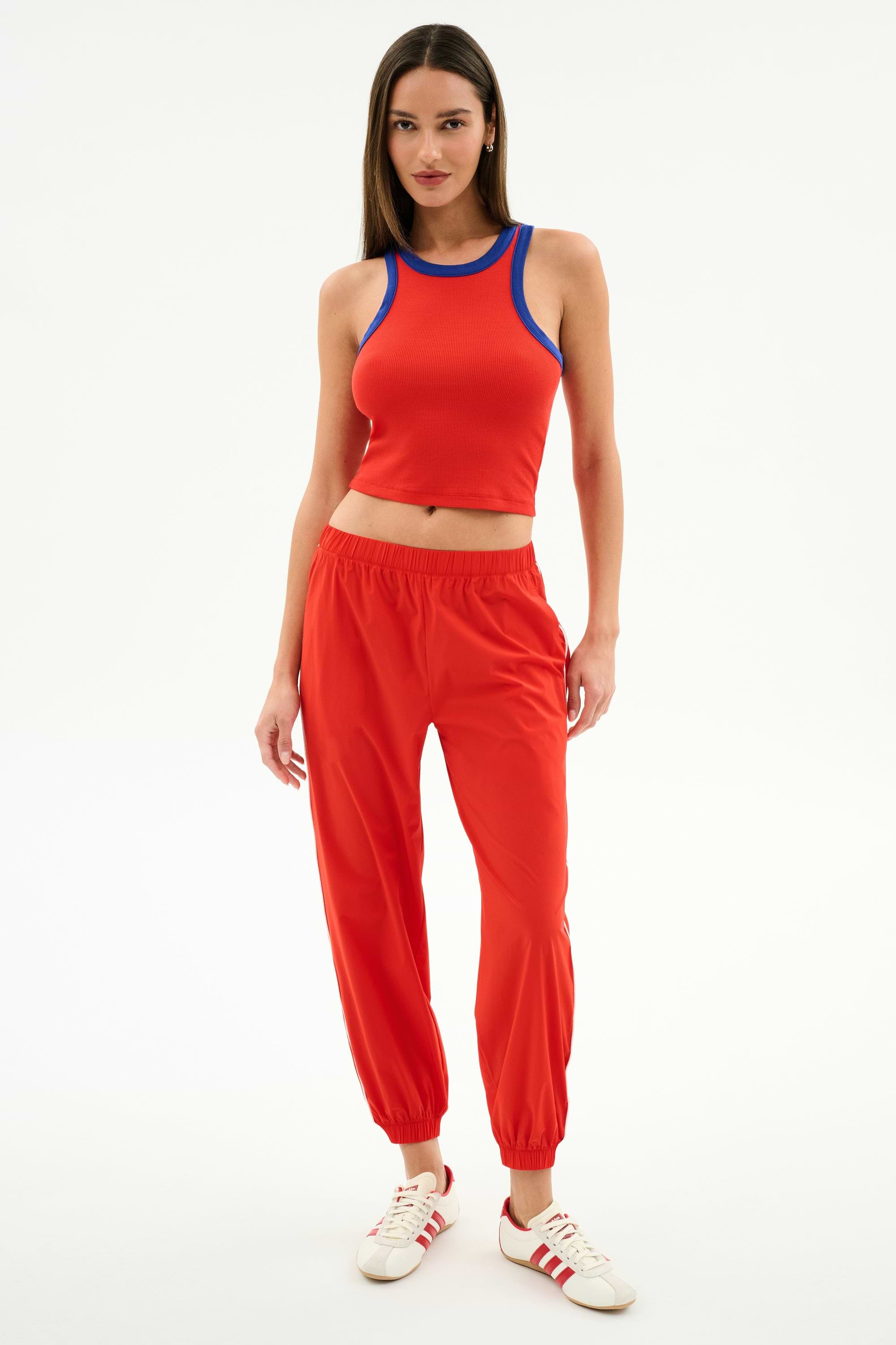 A woman stands against a white background wearing the SPLITS59 Megan Woven Nylon Jogger w/ Piping in Lotus Red/Creme and white sneakers with red stripes.