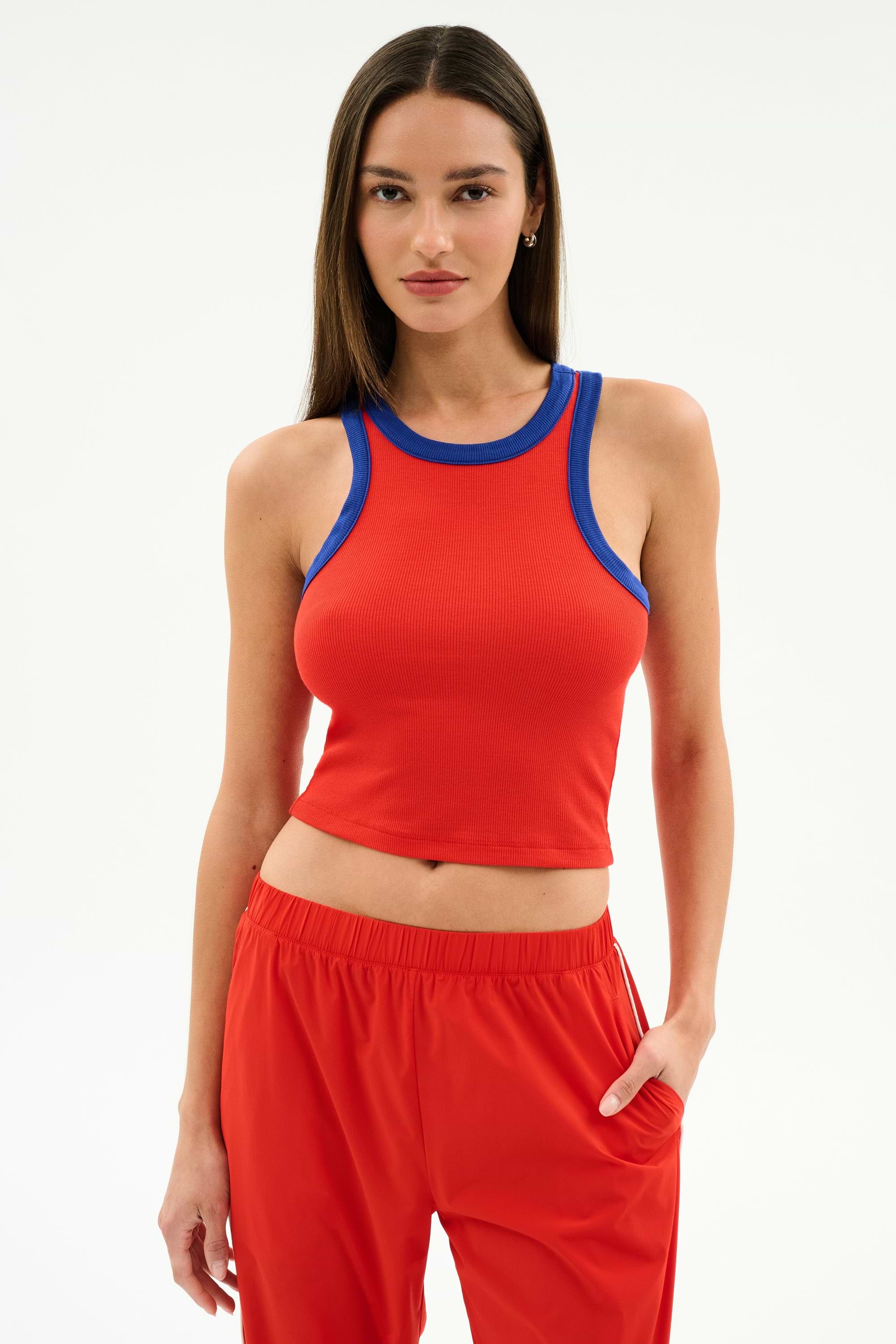 A woman stands against a plain background wearing the SPLITS59 Kiki Rib Crop Tank in Lotus Red/Cobalt with soft baby rib fabric and matching red pants, one hand in her pocket.