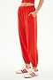 A person wears SPLITS59 Megan Woven Nylon Jogger w/ Piping in Lotus Red/Creme, styled with white sneakers featuring red stripes, and stands with one hand in the pocket against a plain background.
