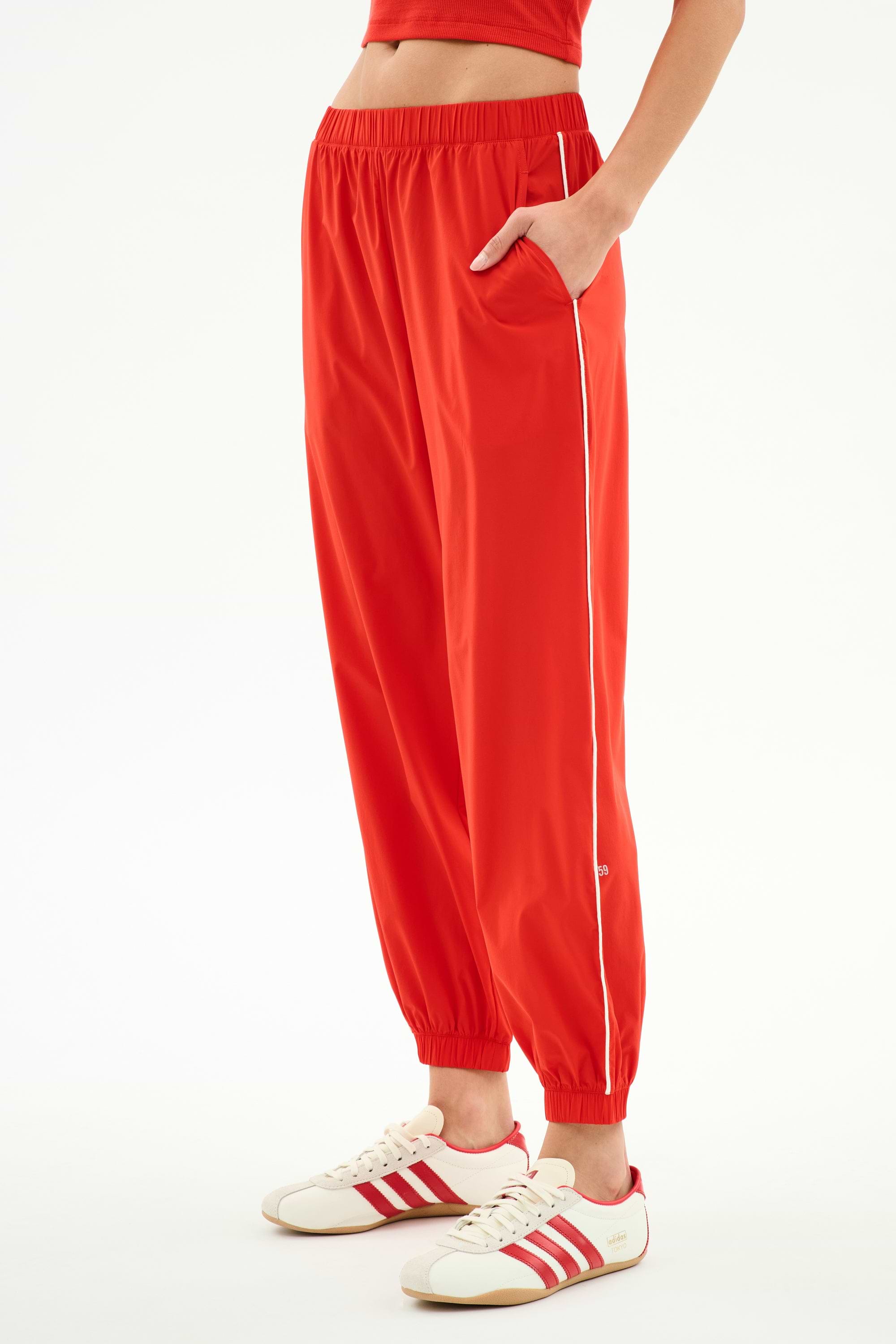 A person wears SPLITS59 Megan Woven Nylon Jogger w/ Piping in Lotus Red/Creme, styled with white sneakers featuring red stripes, and stands with one hand in the pocket against a plain background.