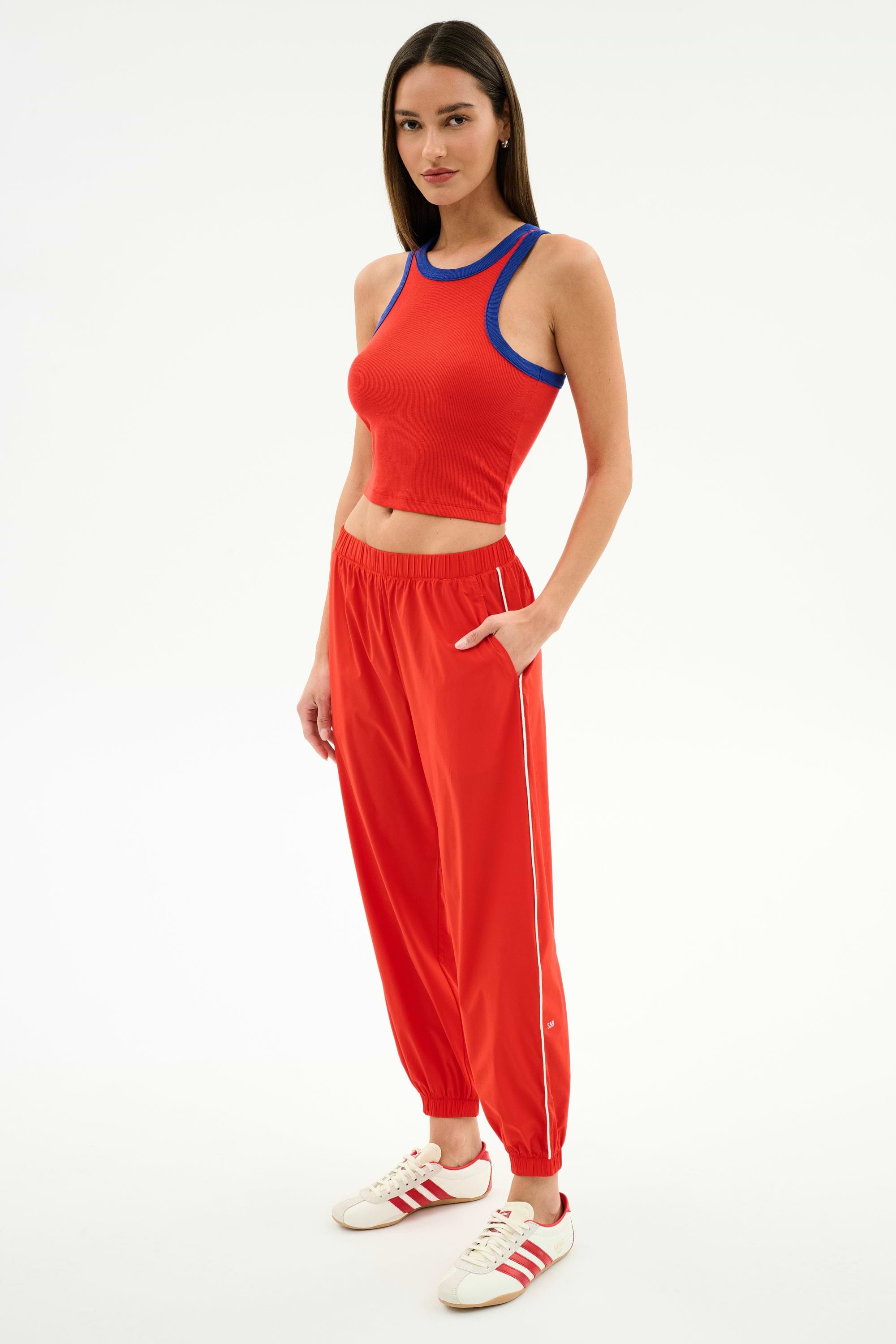 A woman stands against a white background wearing SPLITS59’s Megan Woven Nylon Jogger w/ Piping in Lotus Red/Creme, paired with a red sleeveless crop top and white sneakers with red stripes.