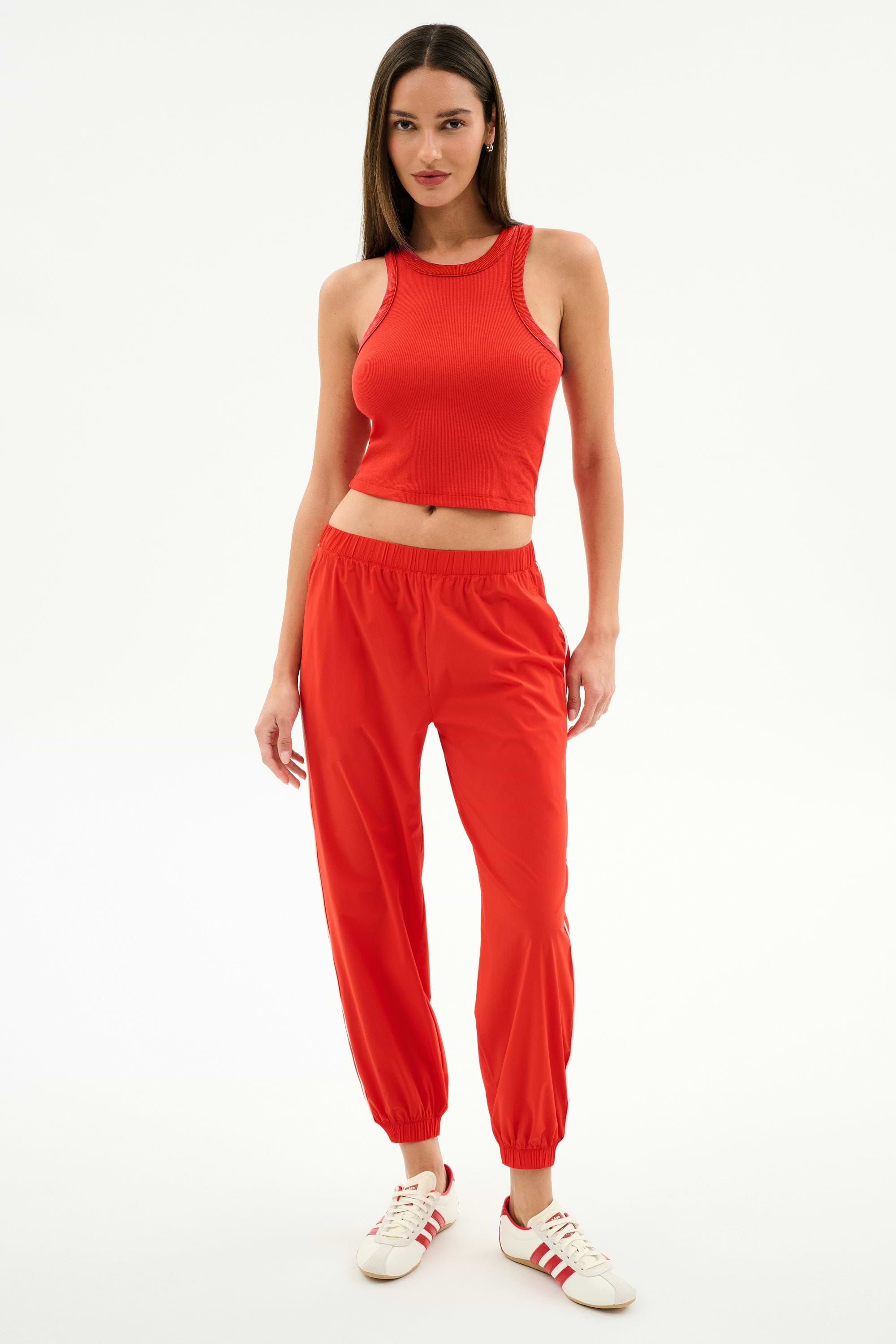 A woman stands against a plain background wearing the SPLITS59 Kiki Rib Crop Tank in Lotus Red, paired with red jogger pants and white sneakers with red stripes—ideal for gym sessions or casual outings.