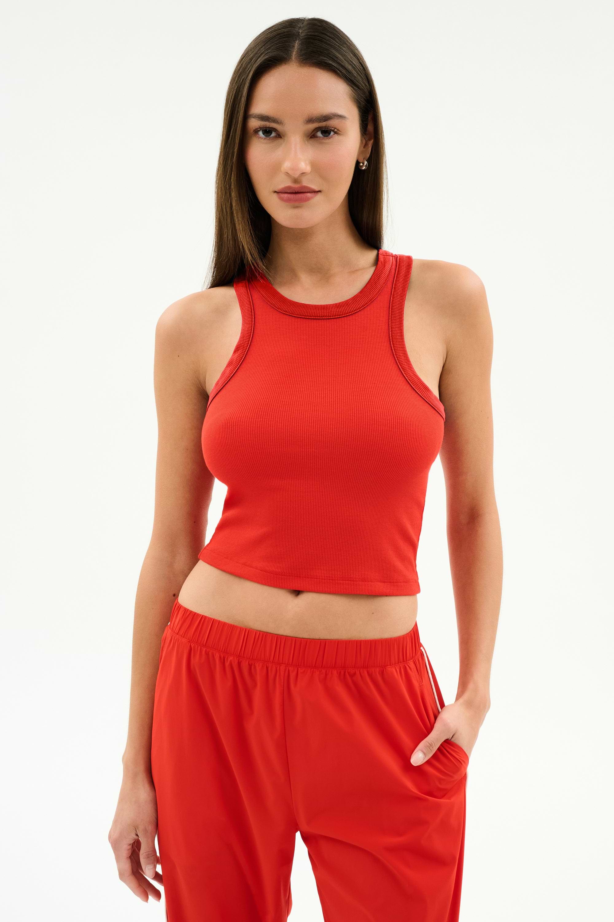 A person stands against a plain background wearing the SPLITS59 Kiki Rib Crop Tank in Lotus Red with matching red pants, one hand in a pocket—ideal for gym sessions or casual outings.