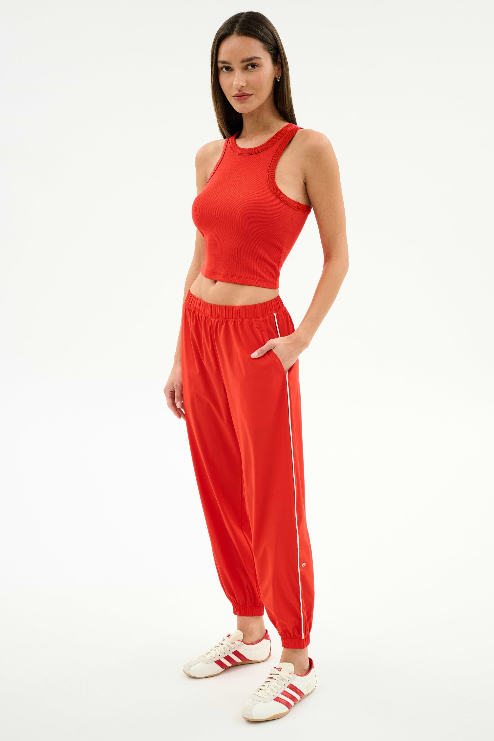 Woman wearing the SPLITS59 Kiki Rib Crop Tank in Lotus Red, paired with red joggers and white Adidas sneakers, standing against a white background. A chic sporty look for street style or gym sessions.