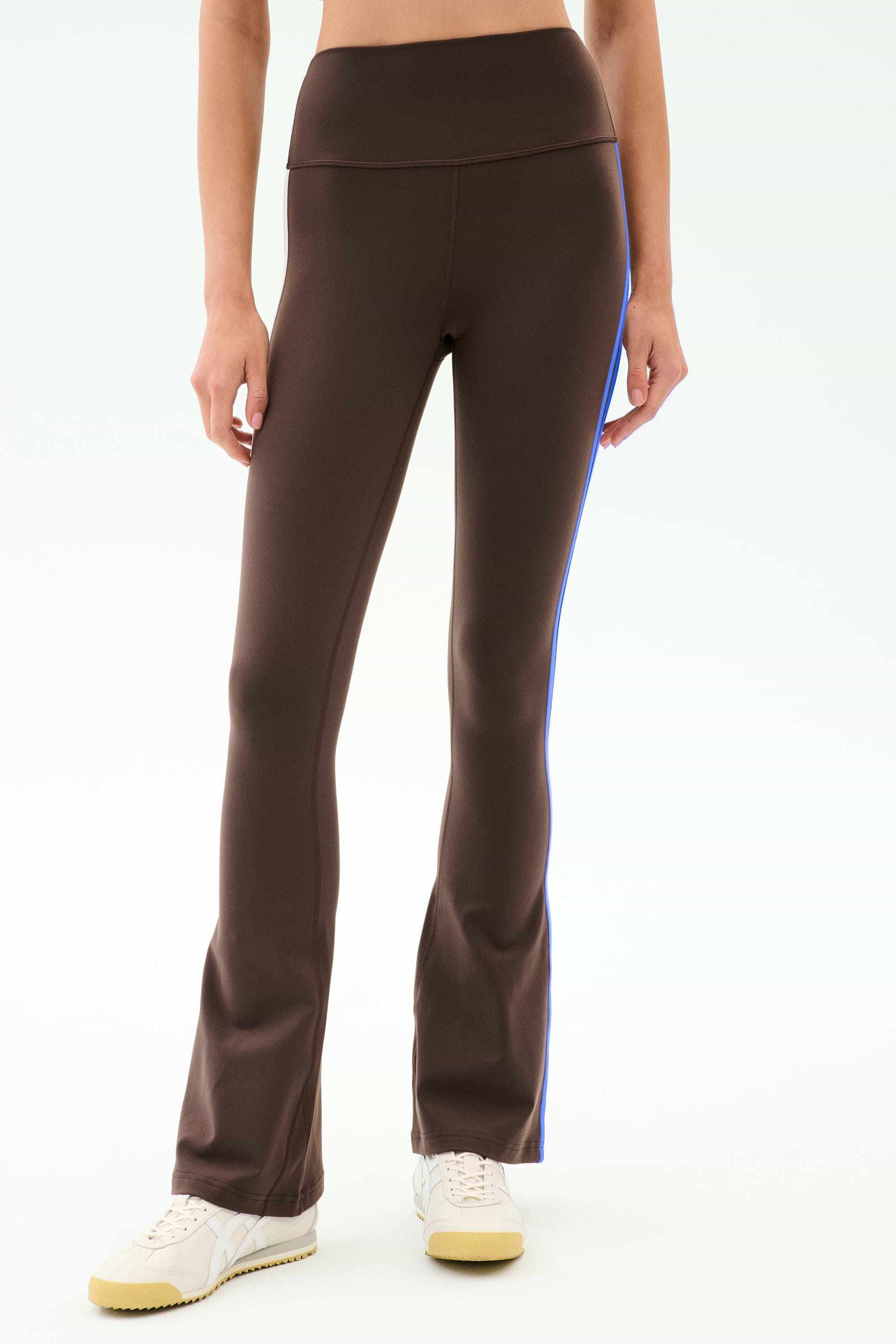 A model is shown from waist to feet wearing SPLITS59 Raquel High Waist Supplex Flare in Dark Chocolate/Classic Blue, paired with white sneakers and a crop top. The 4-way stretch supplex fabric ensures comfort for workouts or all-day wear.