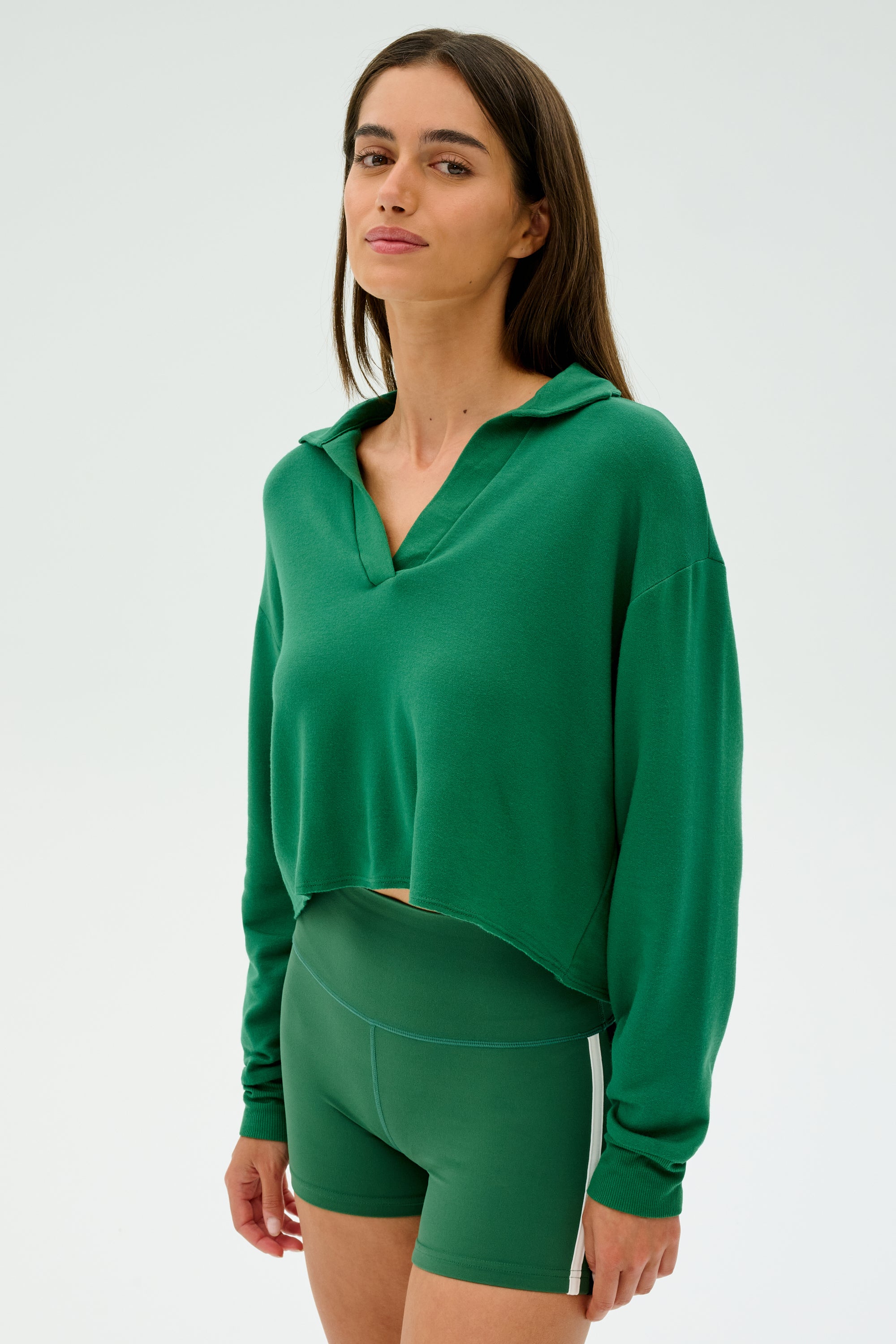 A person wearing the Joan Fleece Polo Sweatshirt in Arugula from SPLITS59 and shorts stands confidently against a plain backdrop, exuding casual elegance in stretch modal fabric.