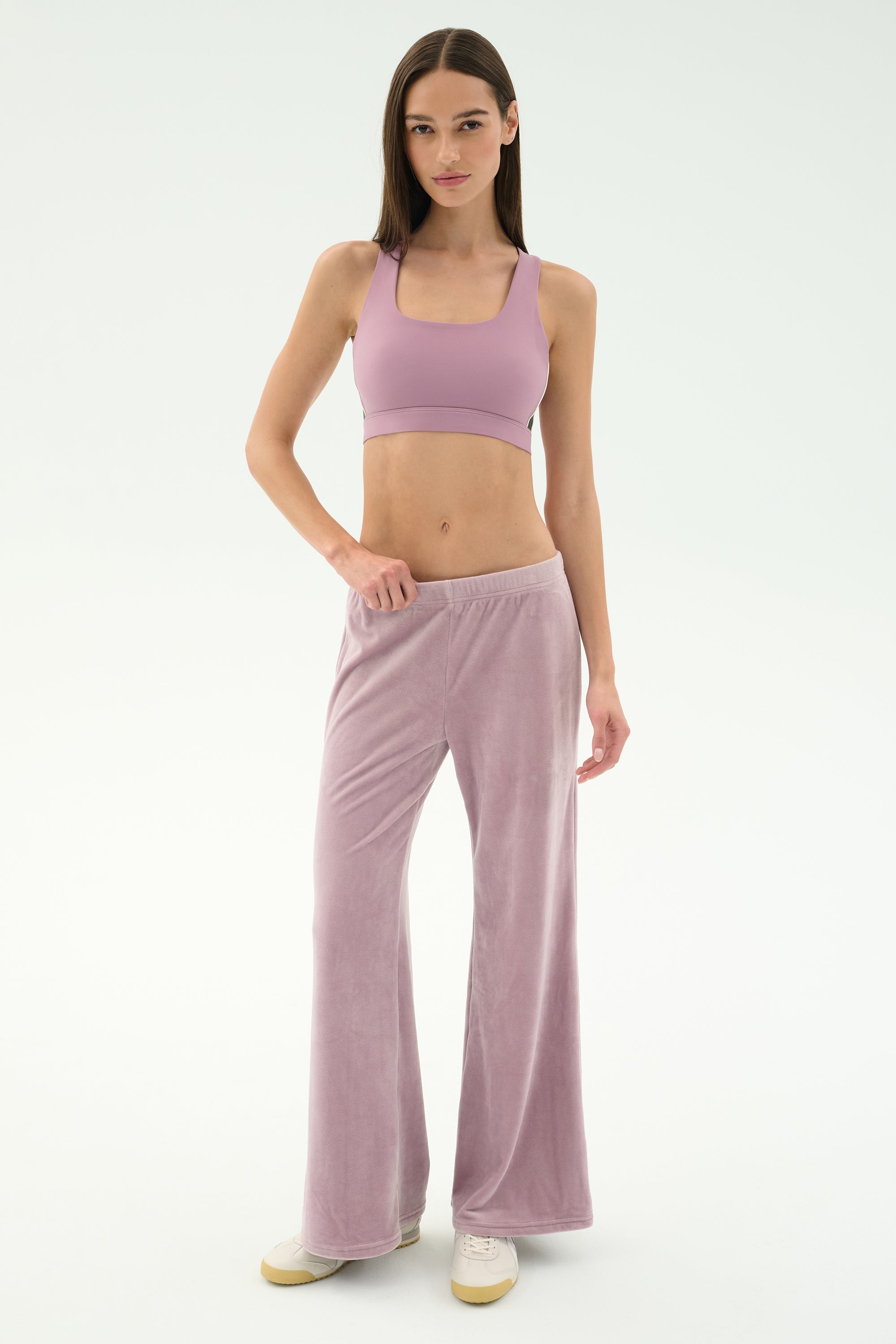 A woman stands in a studio wearing the SPLITS59 Joy Velour Pant in Blush, a mauve sports bra, and white sneakers against a plain white background.