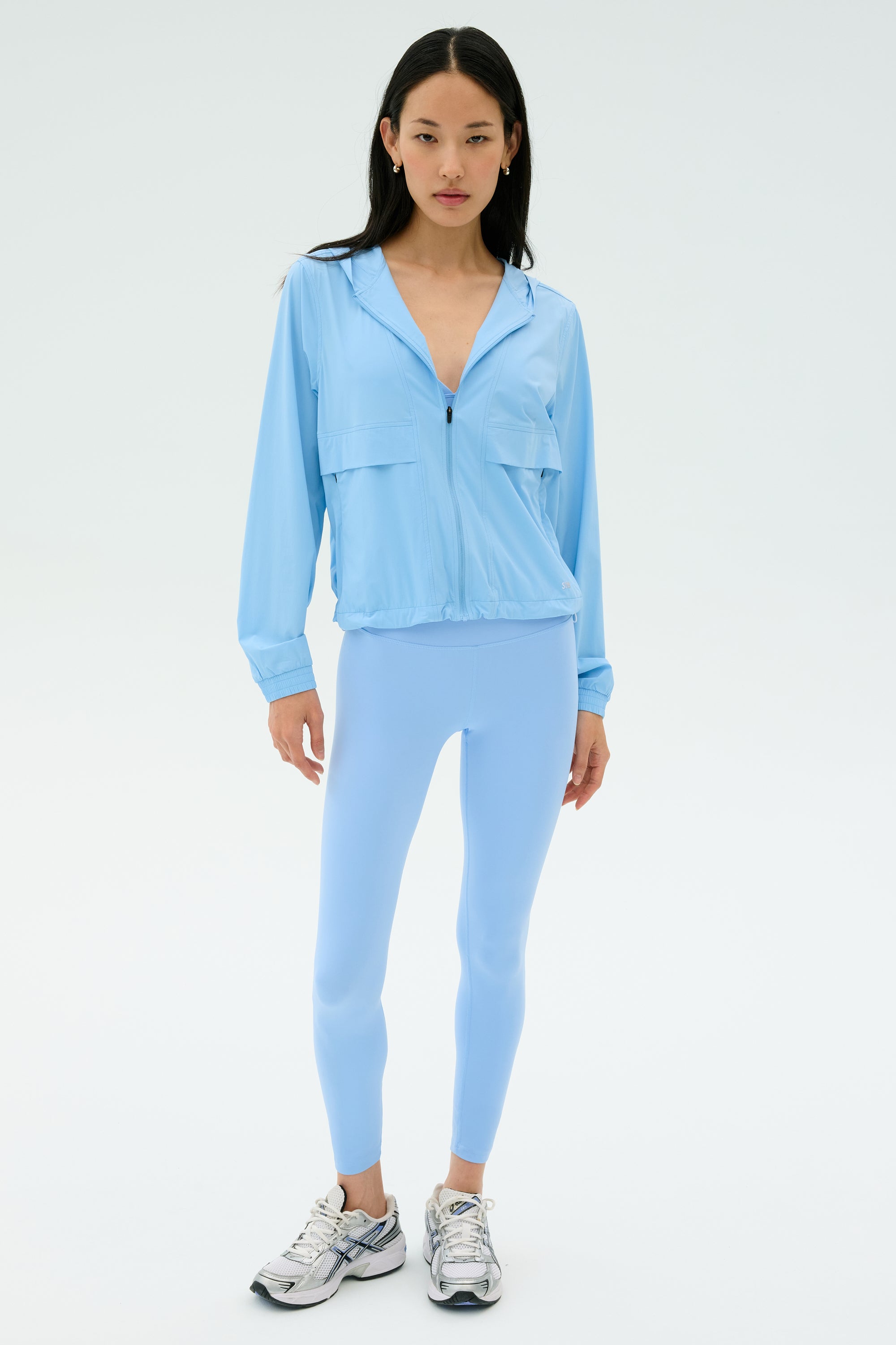 A woman stands against a plain background wearing the SPLITS59 Megan Woven Nylon Jacket in Skylight, paired with matching blue leggings and silver sneakers.