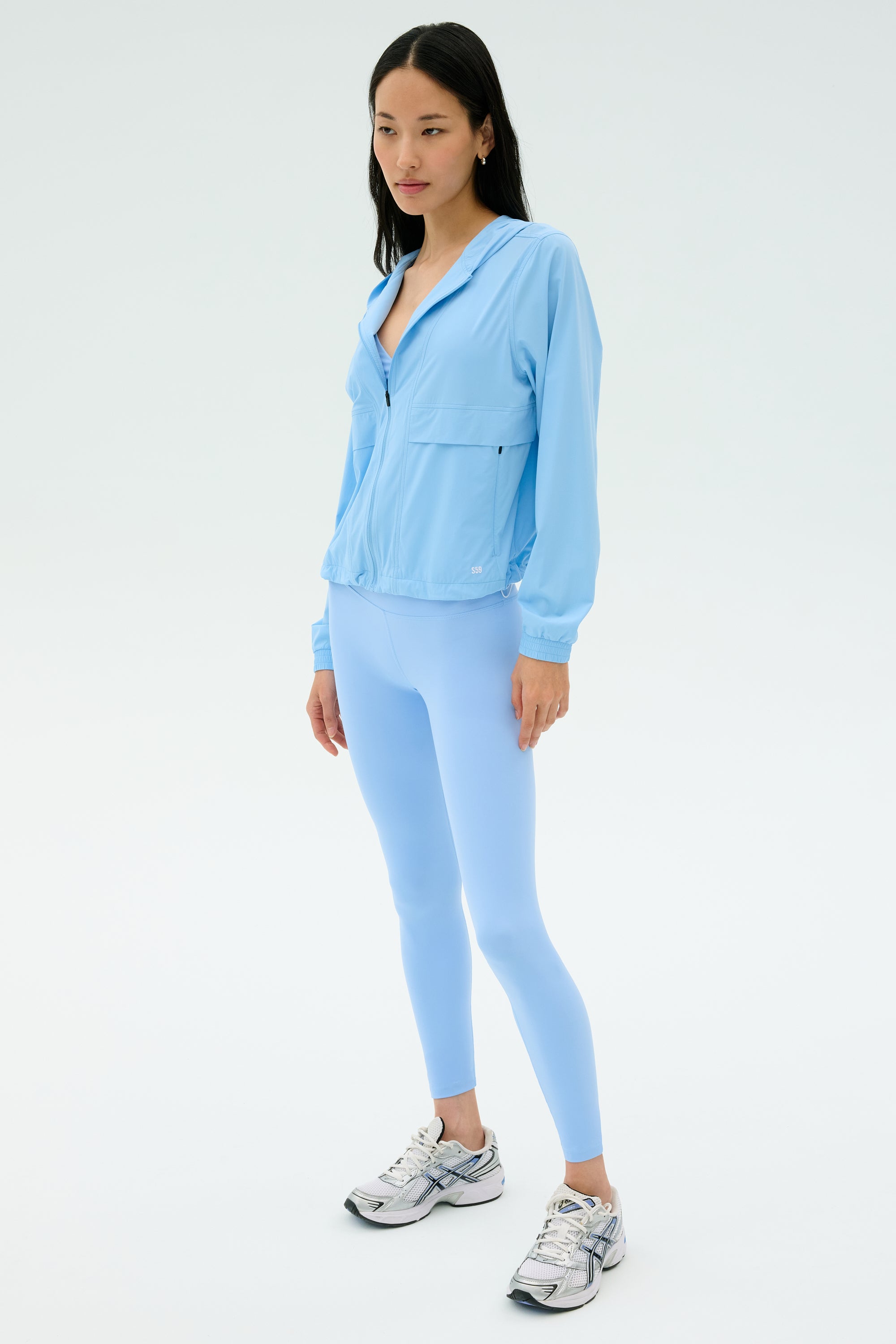 A woman stands against a plain background wearing the SPLITS59 Megan Woven Nylon Jacket in Skylight, paired with matching spandex leggings and silver athletic shoes.