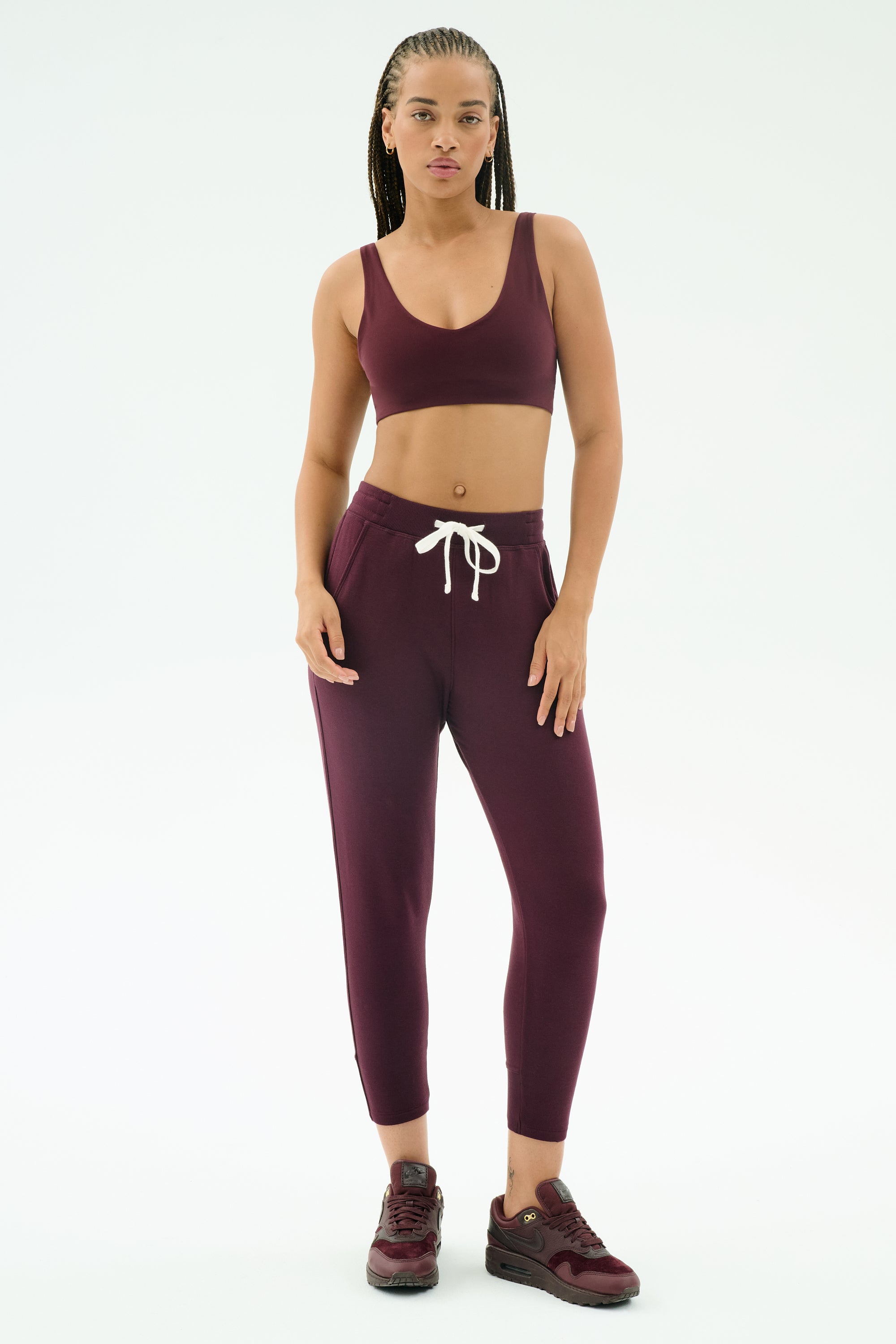 A woman stands against a white background wearing SPLITS59's Reena Fleece 7/8 Sweatpant in Dark Cherry, paired with a matching sports bra and burgundy sneakers.