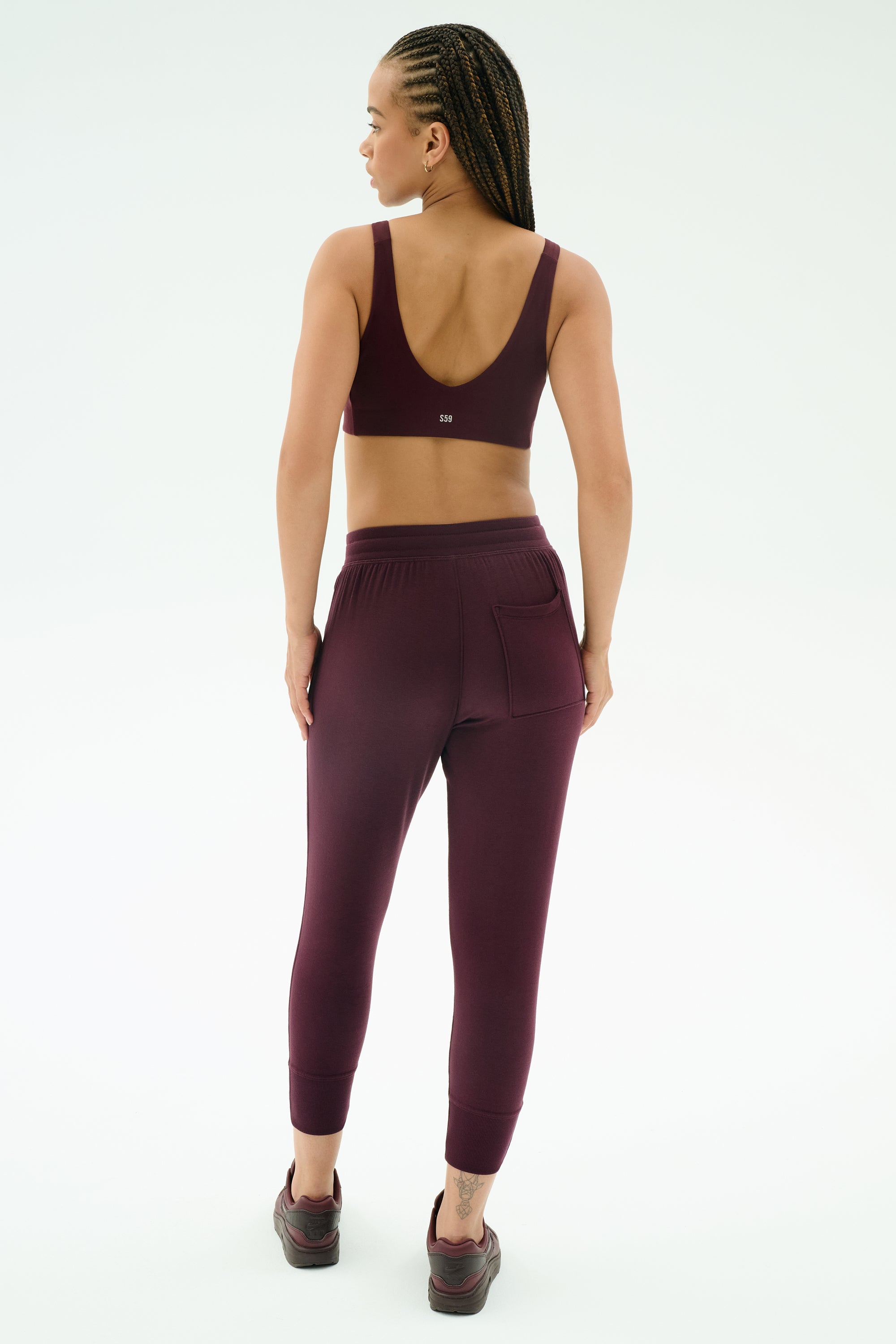 A person wearing SPLITS59's Reena Fleece 7/8 Sweatpant in Dark Cherry stands facing away, highlighting the back details of the soft, stretchy joggers against a plain white background.