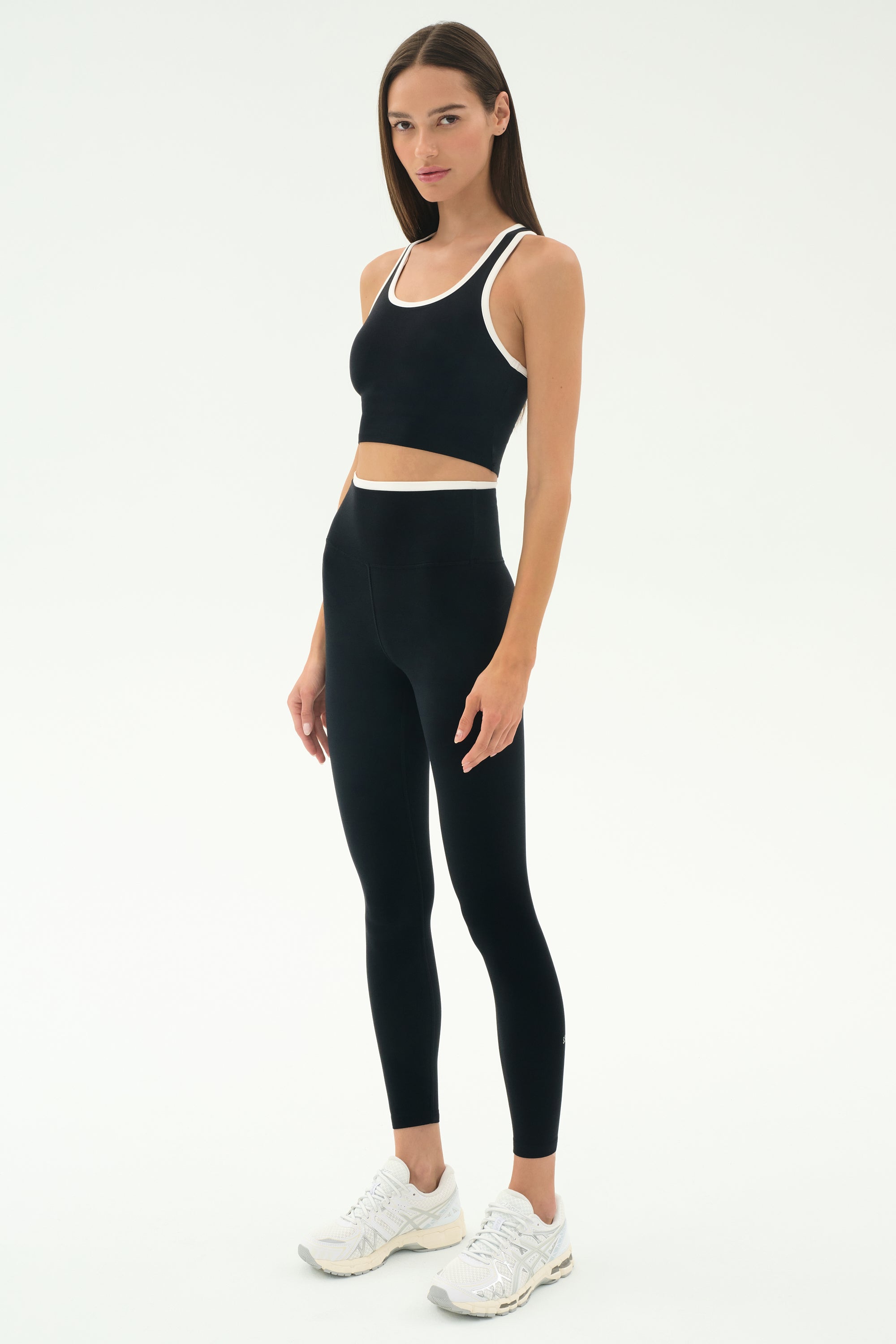 A woman stands against a plain background wearing the SPLITS59 Dual High Waist Airweight 7/8 leggings in Black/White, paired with a black sports bra and white athletic shoes.