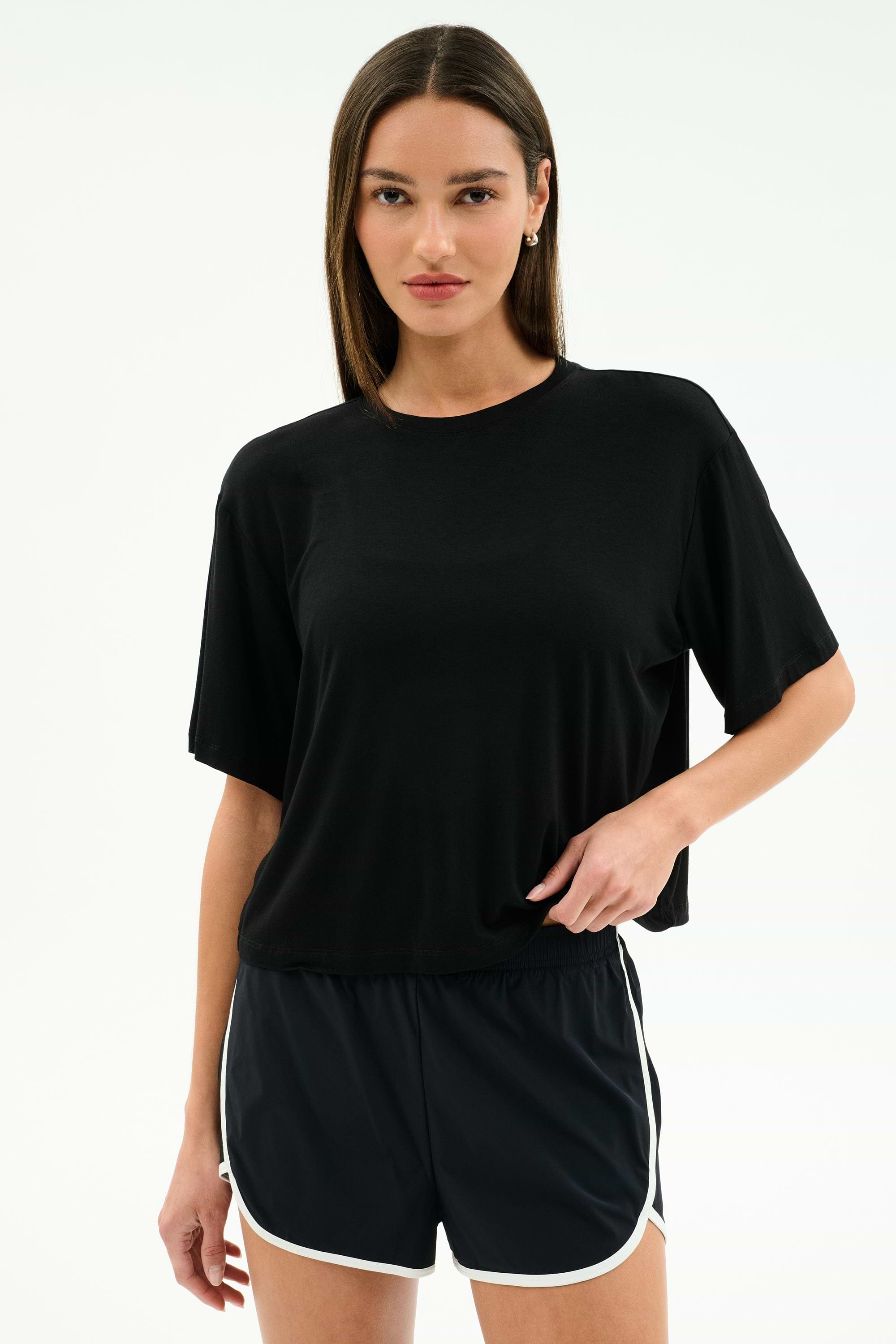 A woman stands against a plain background wearing the SPLITS59 Djuna Oversized Jersey Tee in black and black athletic shorts with white trim, perfect for gym workouts.