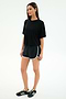 A woman stands against a plain background wearing the SPLITS59 Djuna Oversized Jersey Tee in black, black athletic shorts with white trim, and black Adidas sneakers—an effortless gym-ready look.