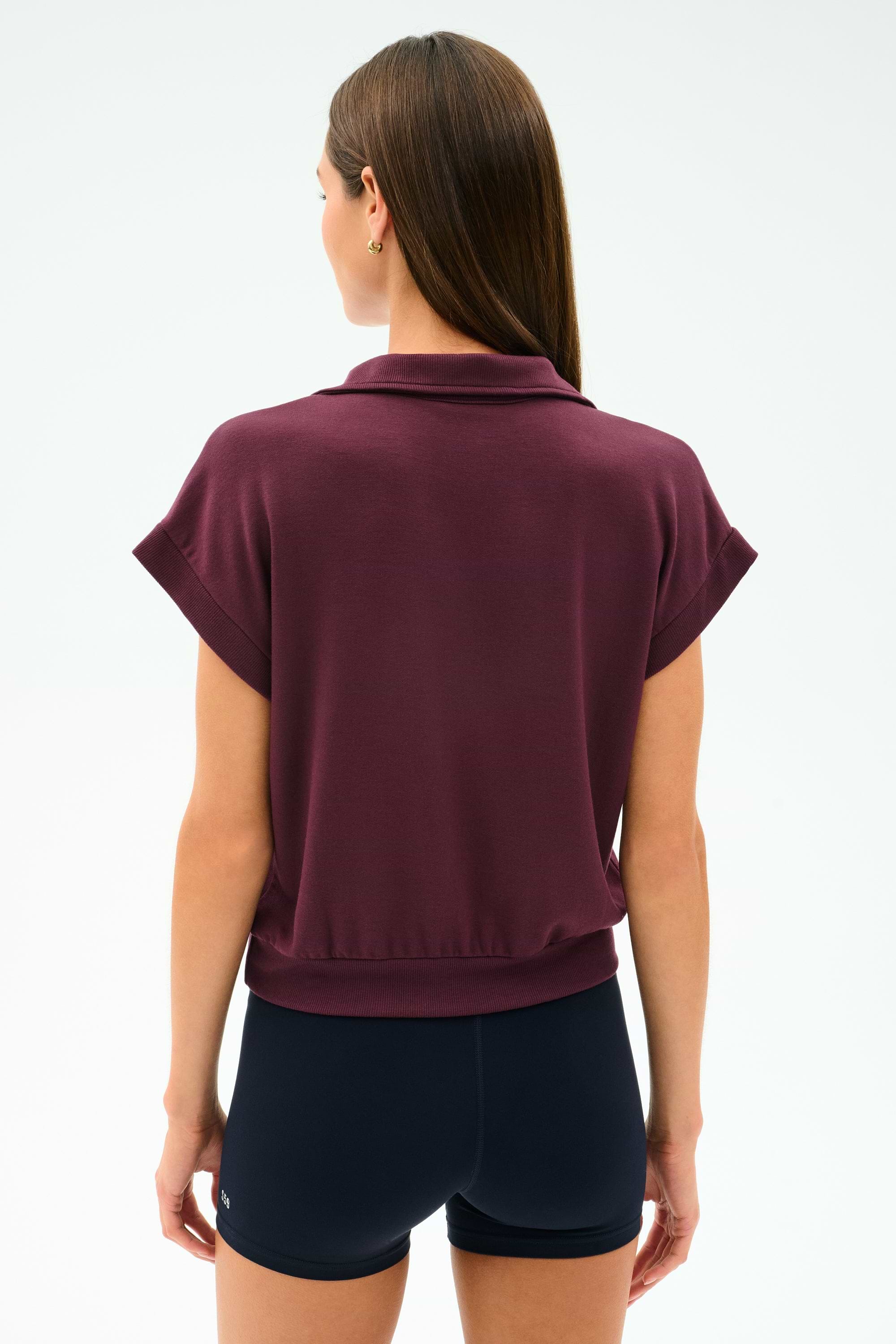 A woman with straight brown hair is seen from the back wearing the SPLITS59 Evie Fleece Sleeveless Polo in Dark Cherry and black shorts against a plain light background.
