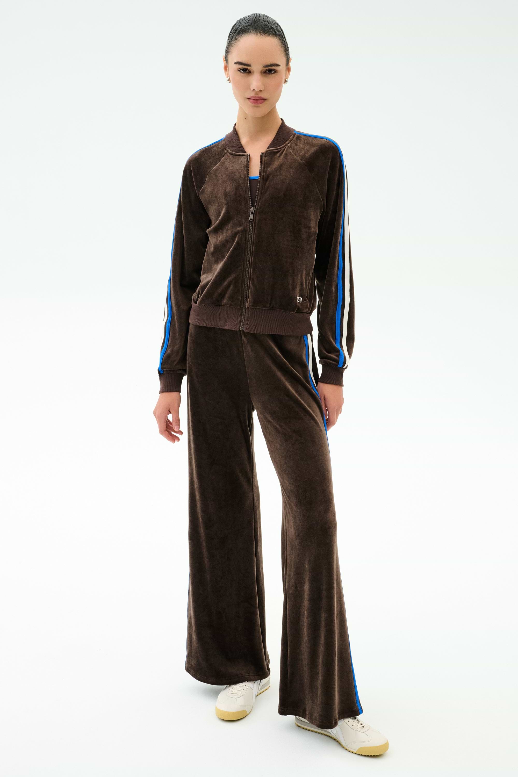 A person stands in SPLITS59 Heritage Velour Pant in Dark Chocolate/Classic Blue featuring blue stripes, wide-leg ankle-length design, paired with white sneakers and a neutral expression against a plain light background.