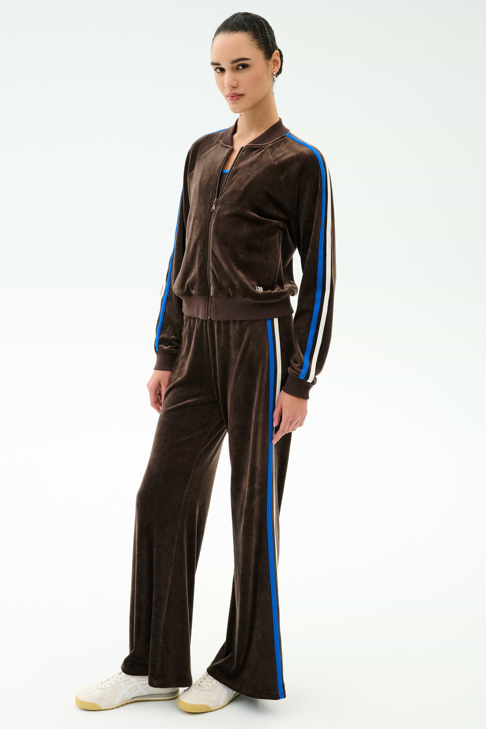 A person wears SPLITS59’s Heritage Velour Pant in Dark Chocolate/Classic Blue, featuring wide-leg comfort and blue and white stripes, paired with white sneakers against a plain light background.