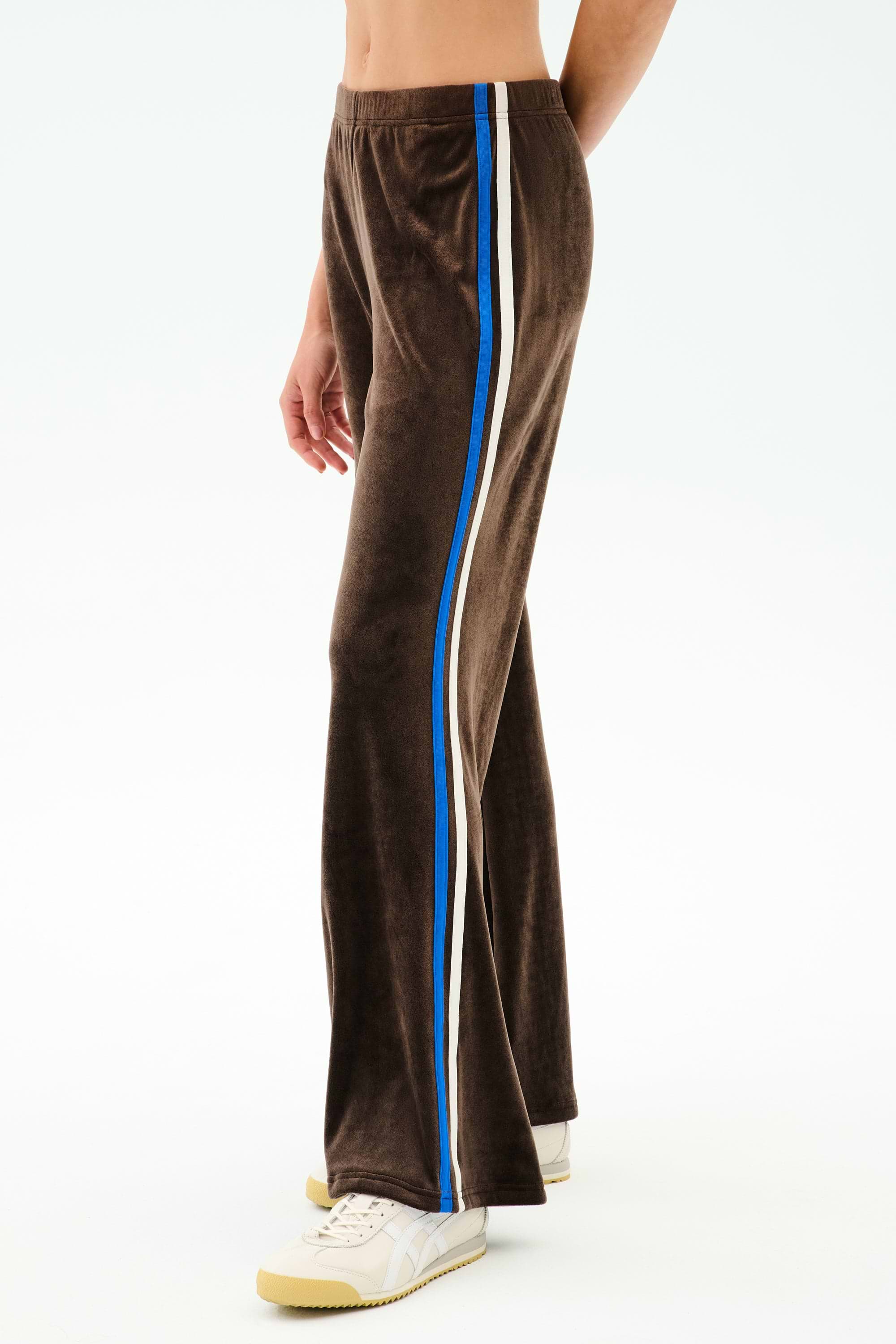 A person wears the SPLITS59 Heritage Velour Pant in Dark Chocolate/Classic Blue with blue and white side stripes, paired with white sneakers and a bare midriff, standing against a plain white background.