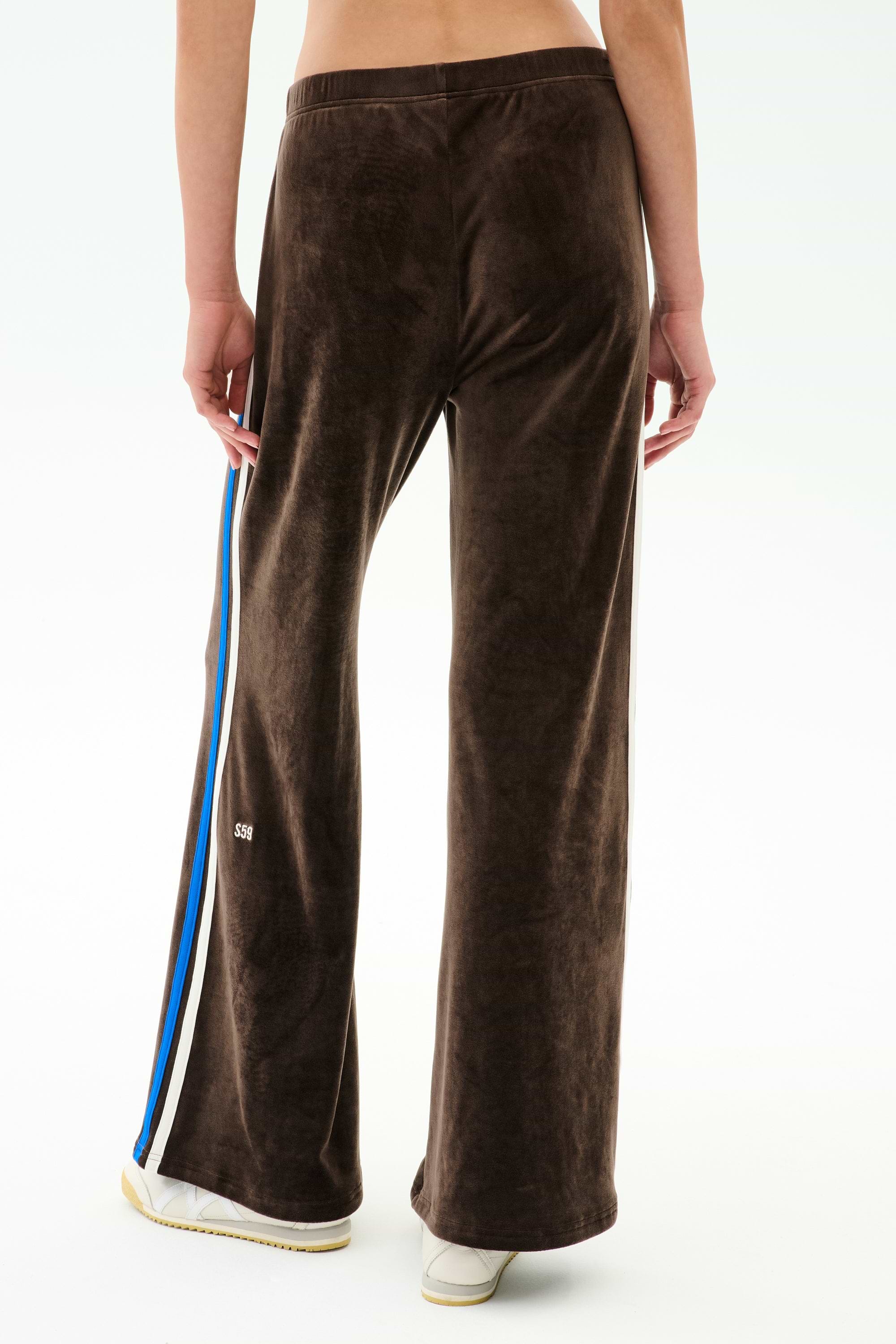 A person models the SPLITS59 Heritage Velour Pant in Dark Chocolate/Classic Blue—ankle-length, wide-leg silhouette with blue and white side stripes—paired with white sneakers, shown from the back against a plain background.