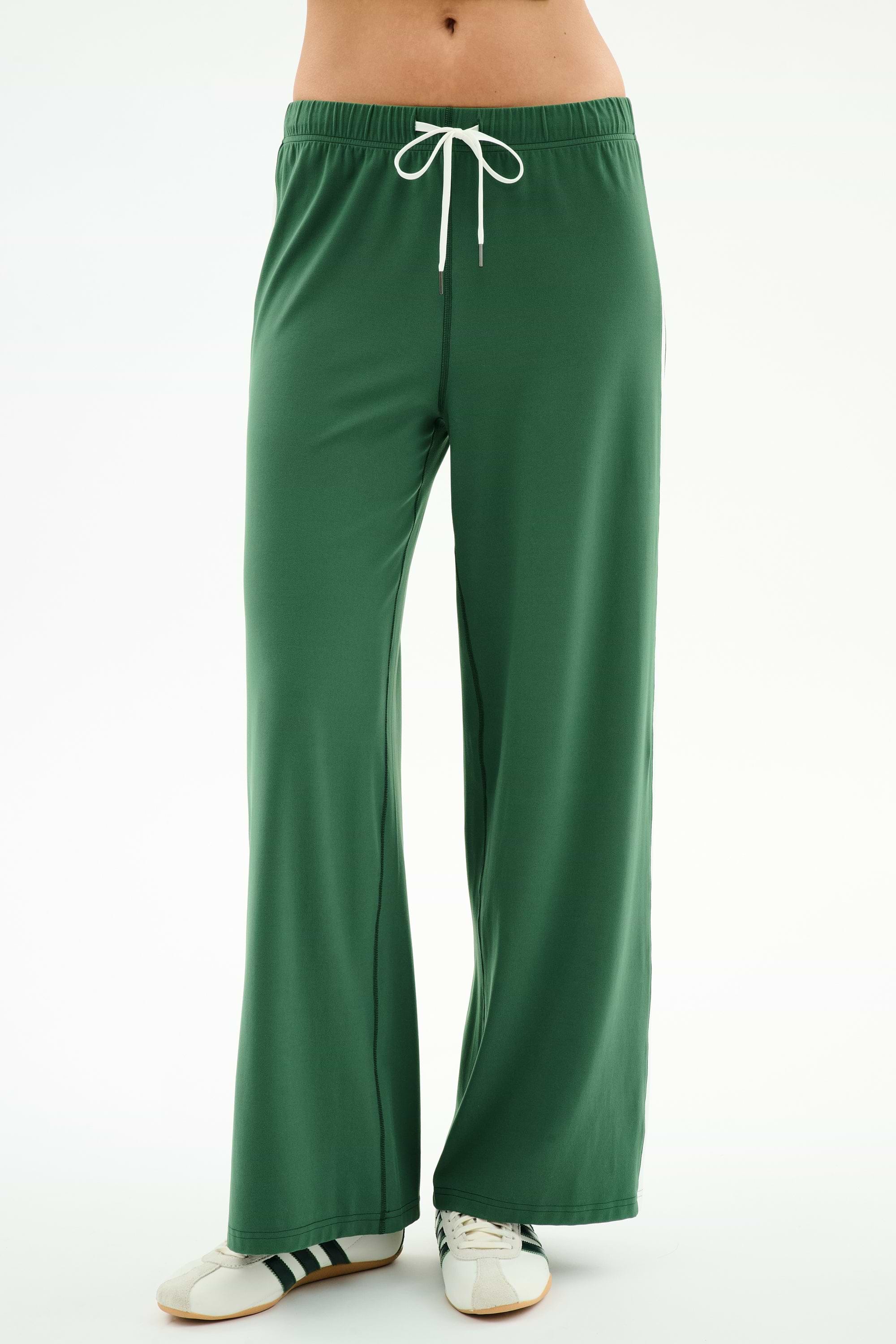 A person wearing SPLITS59 Willow Airweight Wide Leg pants in Arugula/White and white sneakers with black stripes stands against a plain white background.