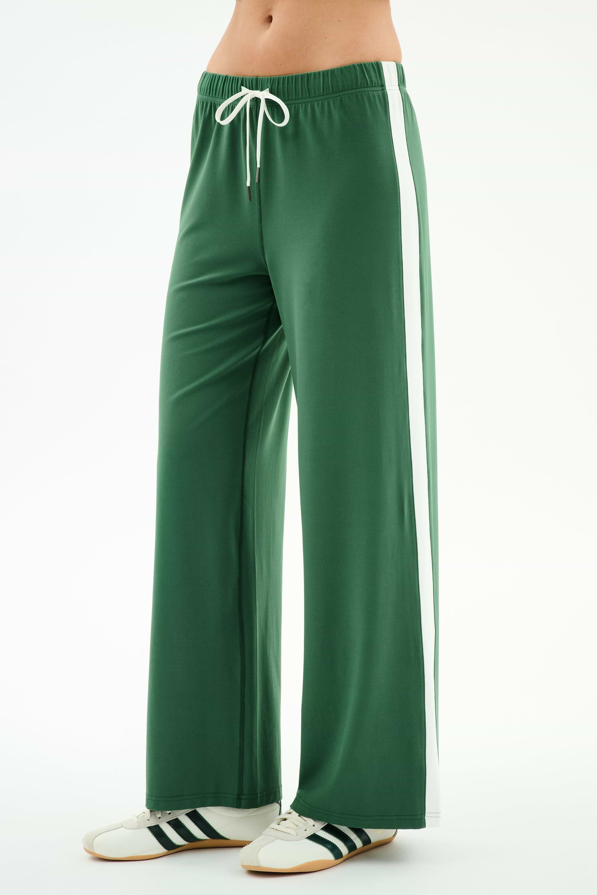 A person wears SPLITS59 Willow Airweight Wide Leg pants in Arugula/White, featuring white side stripes and a drawstring waist, paired with white sneakers with black stripes, standing against a plain background.