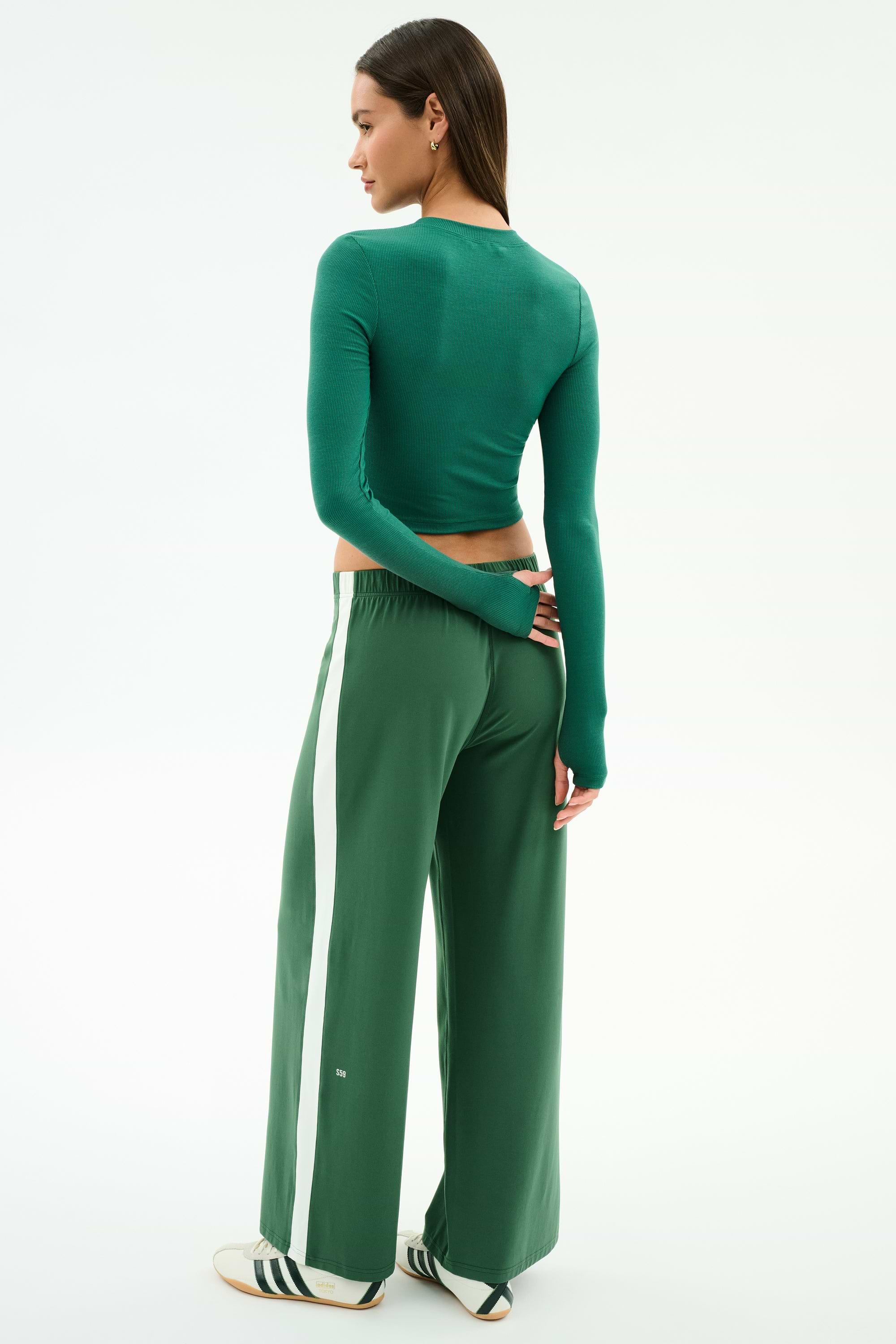 A woman faces away, wearing SPLITS59's Willow Airweight Wide Leg pants in Arugula/White and a fitted green spandex long-sleeve top, paired with white sneakers featuring black stripes.