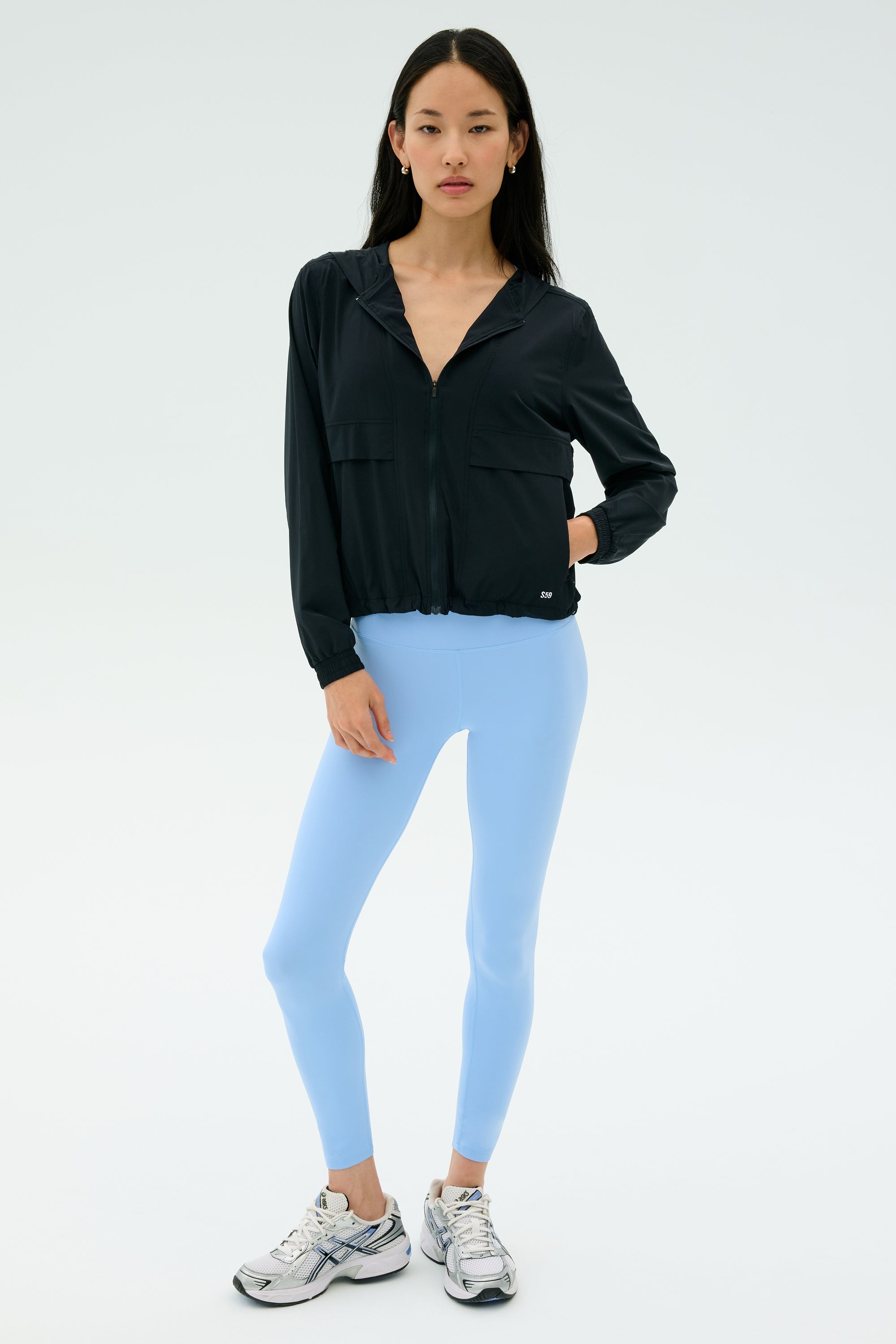 A woman stands against a plain background wearing the SPLITS59 Megan Woven Nylon Jacket in black, paired with light blue spandex leggings and silver athletic shoes.