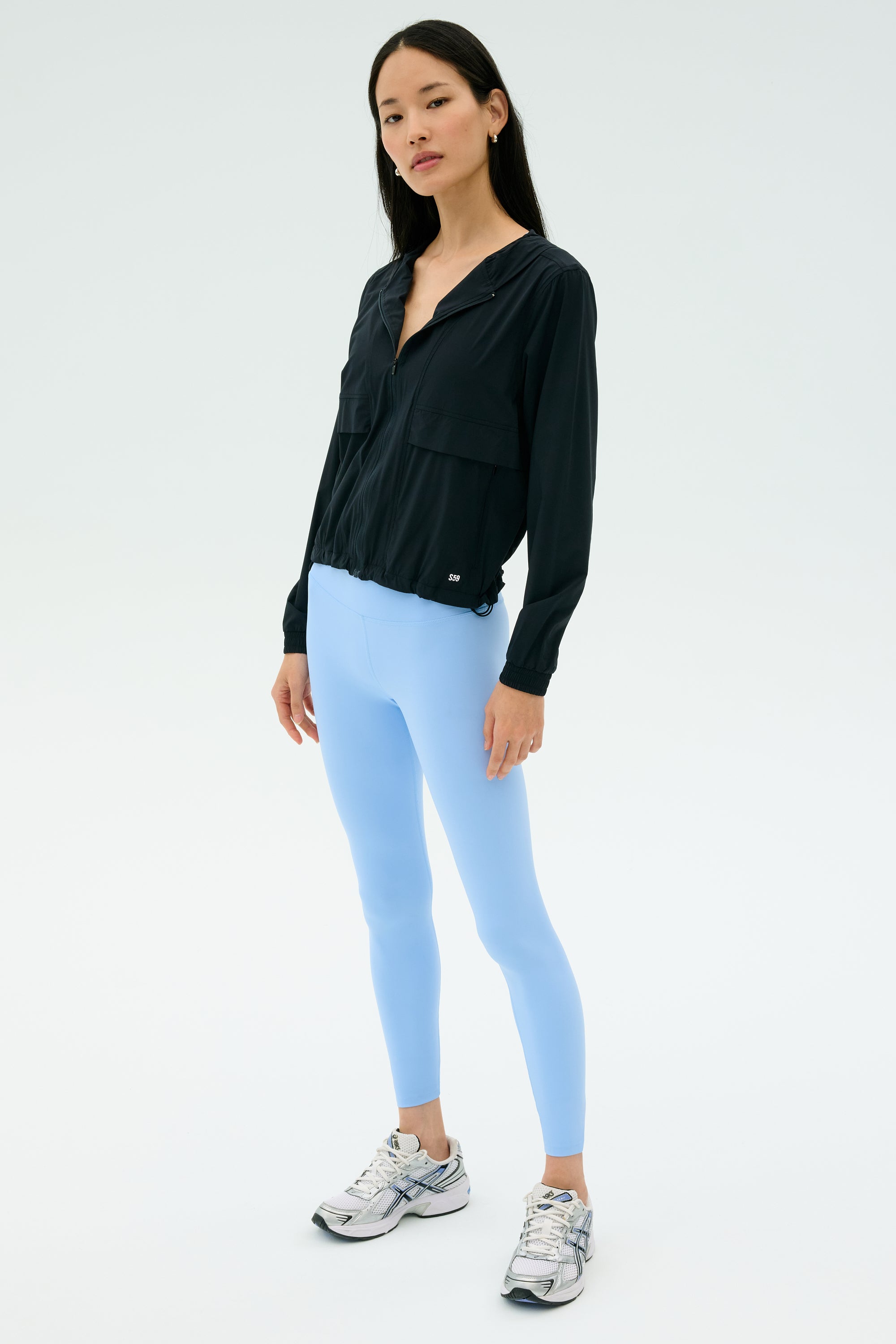 A woman wears the SPLITS59 Megan Woven Nylon Jacket in black, paired with light blue leggings and silver running shoes, set against a plain light background.