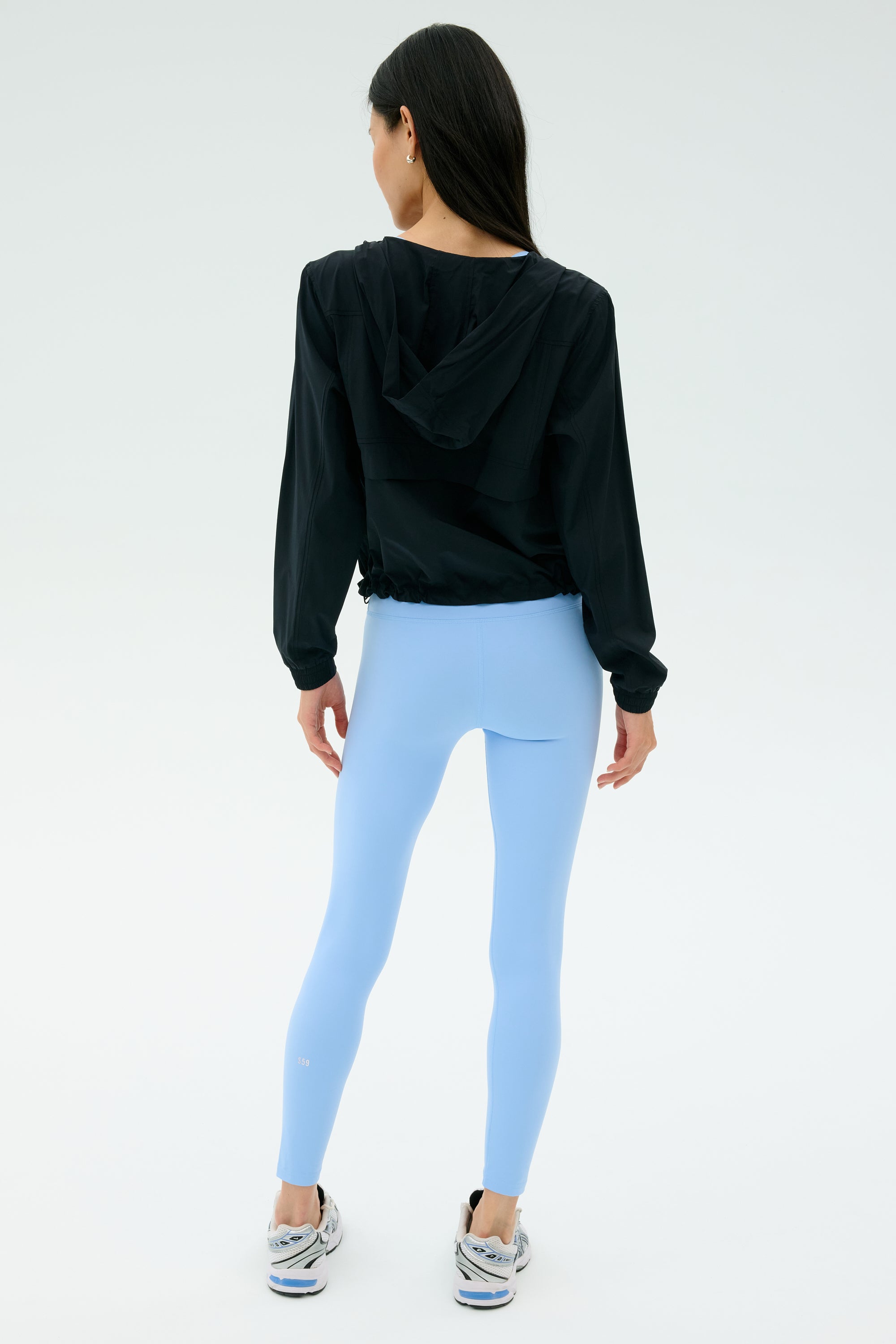 A person stands facing away, wearing the SPLITS59 Megan Woven Nylon Jacket in black, paired with light blue spandex leggings and white sneakers against a plain light background.