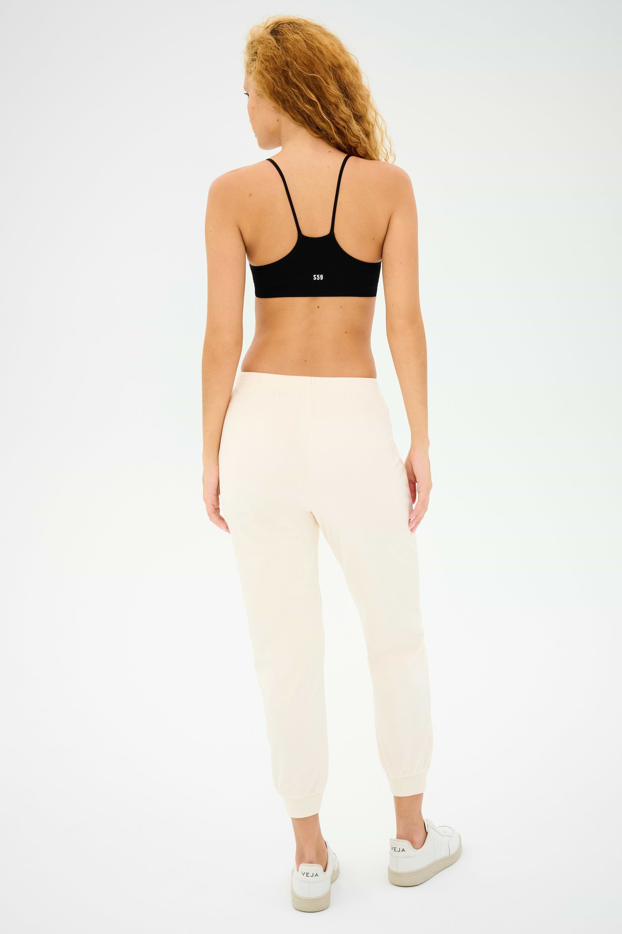 A curly-haired person stands with their back to the camera, poised for gym workouts in a Splits59 Loren Seamless Bra - Black, light joggers, and white sneakers against a plain backdrop.