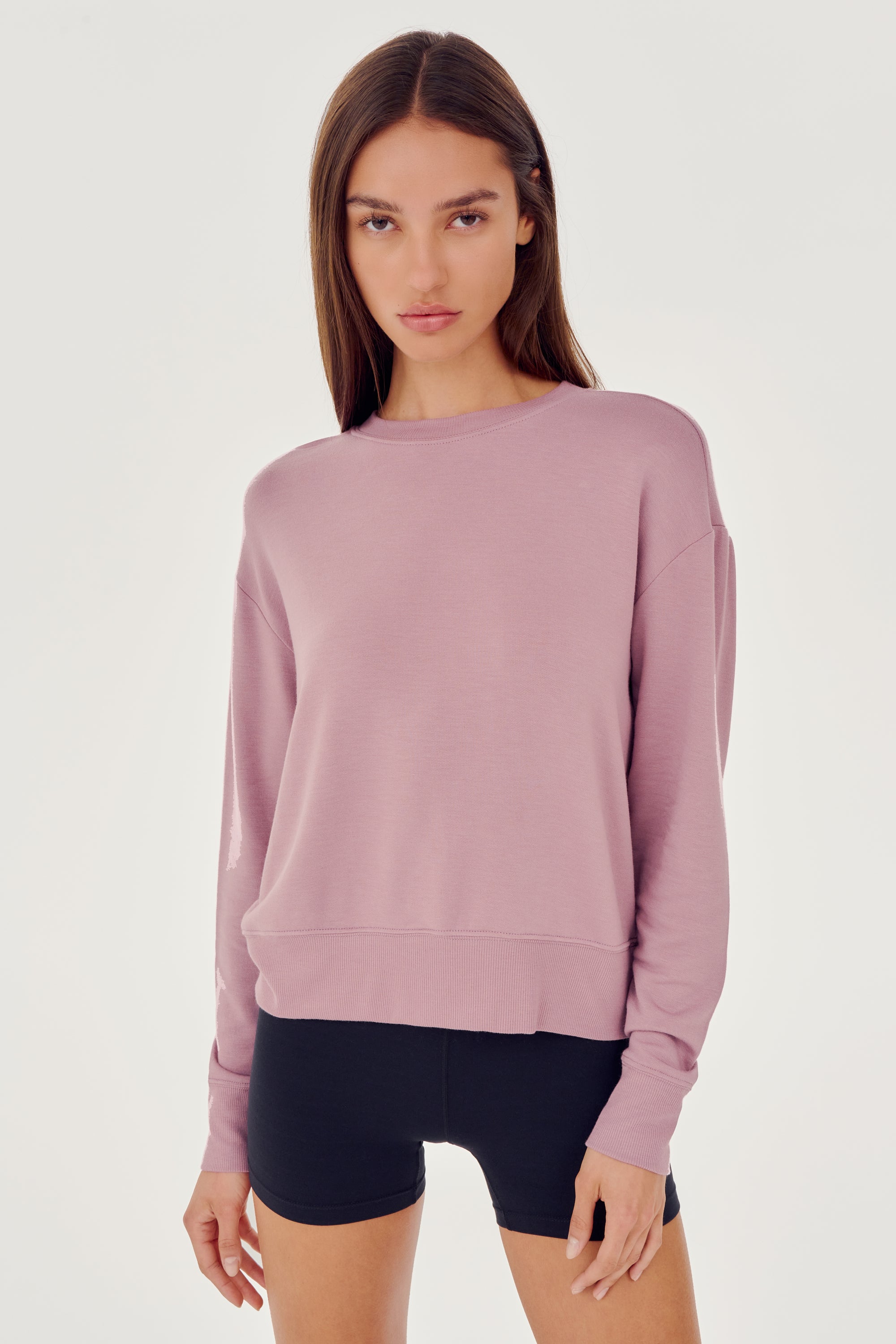 Front view of woman with dark brown hair wearing a light pink crewneck sweatshirt and black shorts 