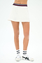 A person models the SPLITS59 Kaia Active Rib Skort in white, featuring a striped waistband, paired with white socks and athletic shoes—ideal for court sports or gym workouts—against a plain white background.
