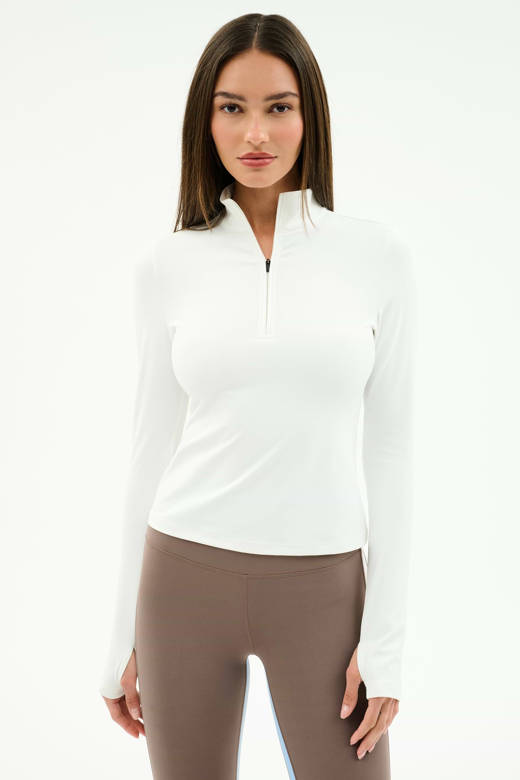 A woman faces forward against a plain white background, wearing the SPLITS59 Tess Rigor Half Zip Waist Length top in white and taupe nylon spandex leggings.