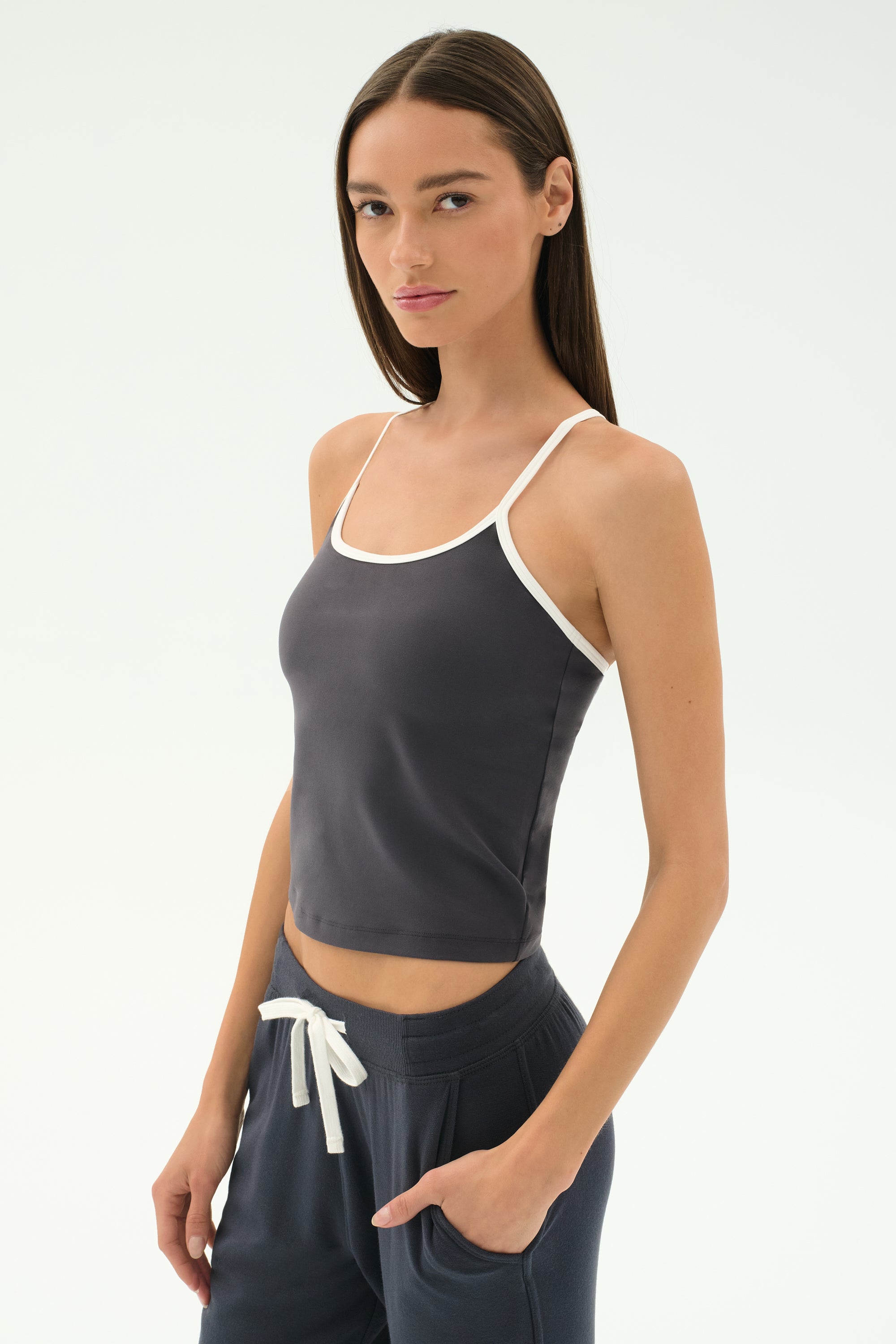 A woman wears the SPLITS59 Airweight Tank in Graphite/White with matching drawstring pants, standing with one hand in her pocket, ready for Pilates or hot yoga against a plain white background.