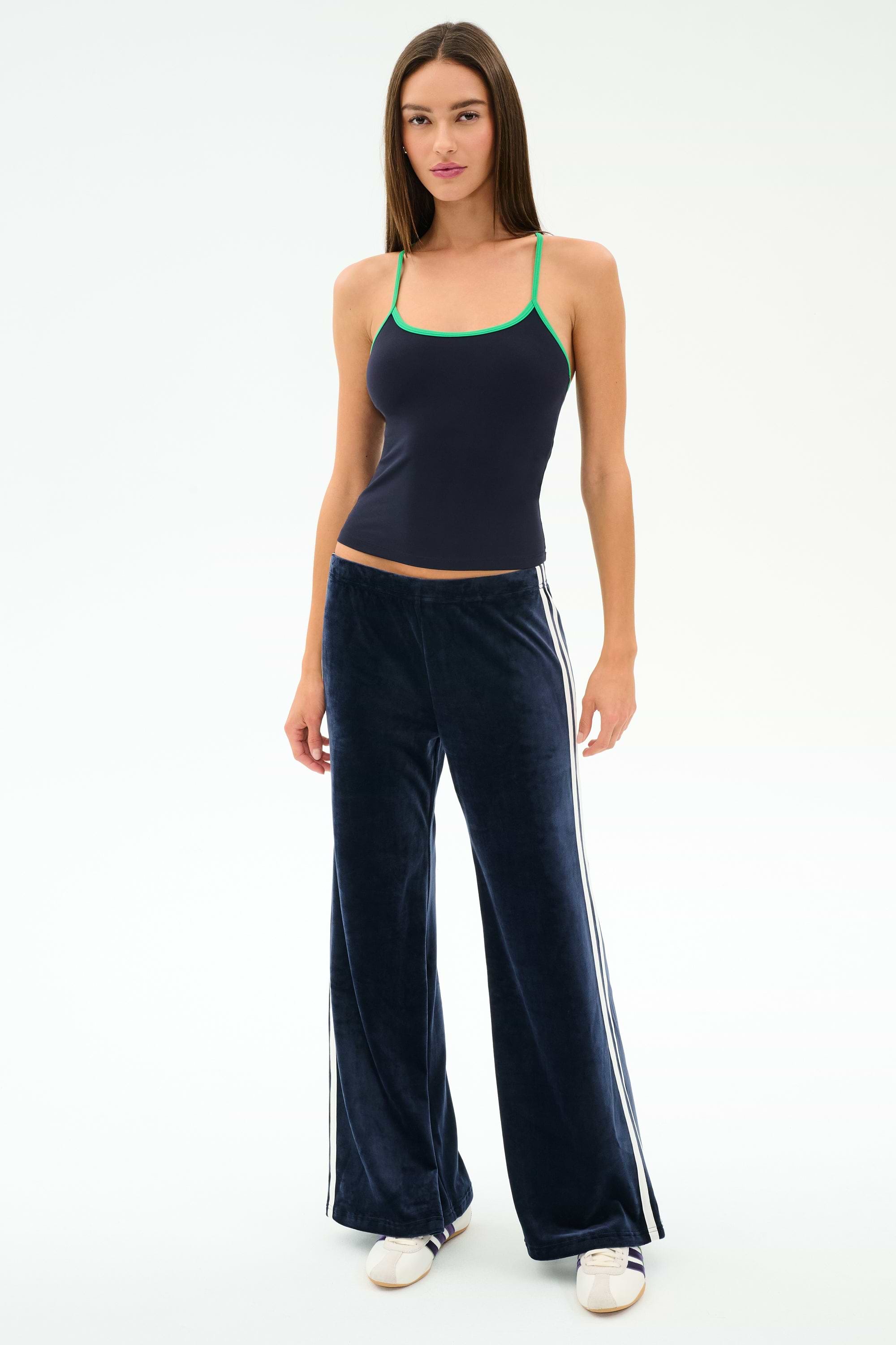 A woman stands against a white background wearing SPLITS59 Heritage Velour Pant in Indigo/White along with a navy camisole with green straps and white sneakers.