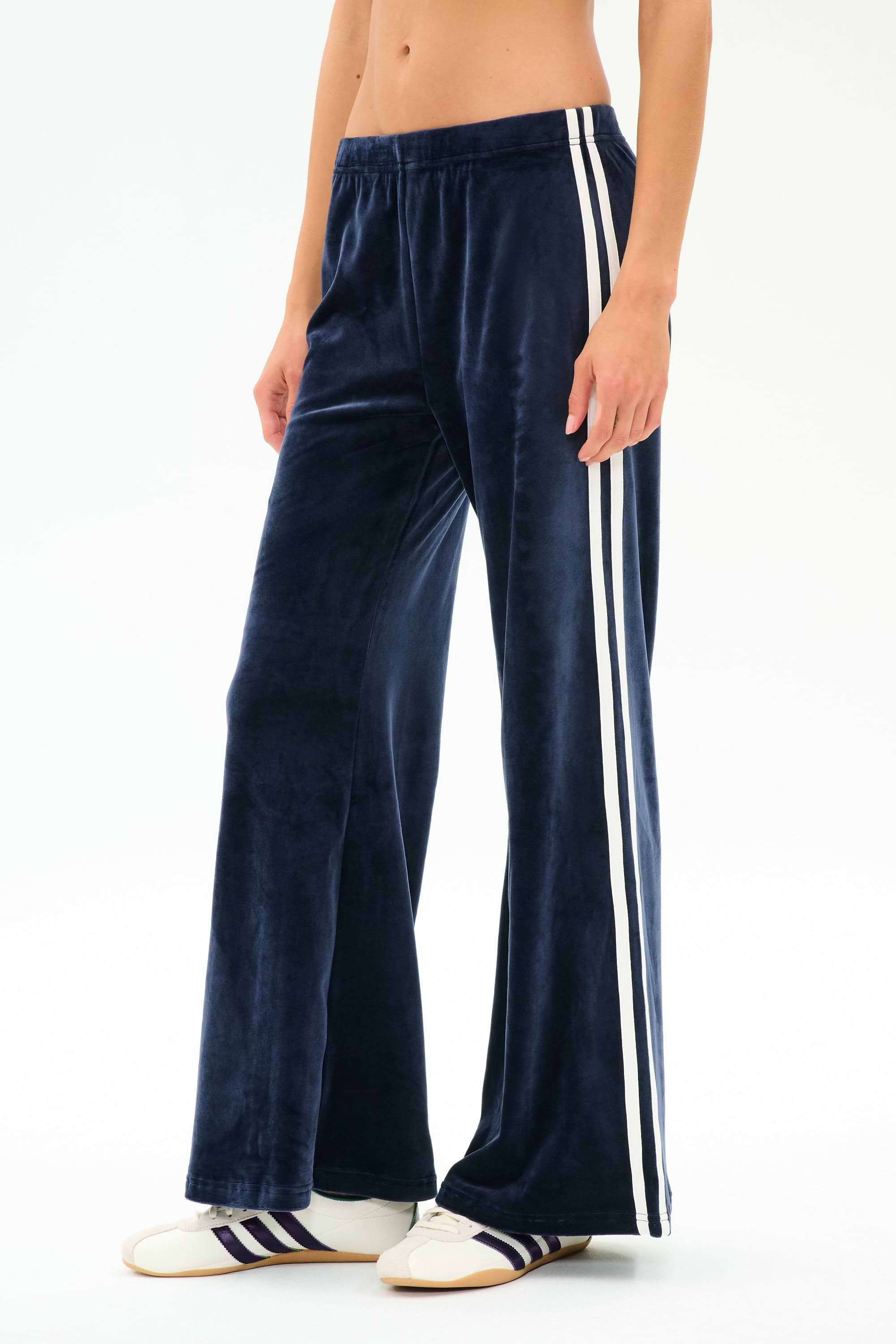 A person wears SPLITS59 Heritage Velour Pant in Indigo/White, featuring white side stripes, paired with white sneakers with navy stripes, standing against a plain light background.