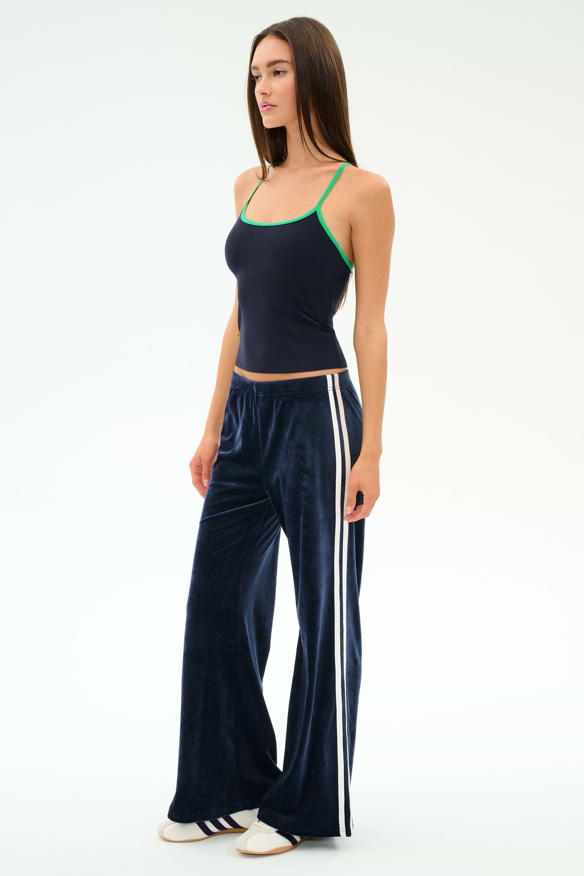 A woman wears SPLITS59’s Heritage Velour Pant in Indigo/White with white side stripes, navy spandex tank top with green trim, and white shoes, standing against a plain light background.