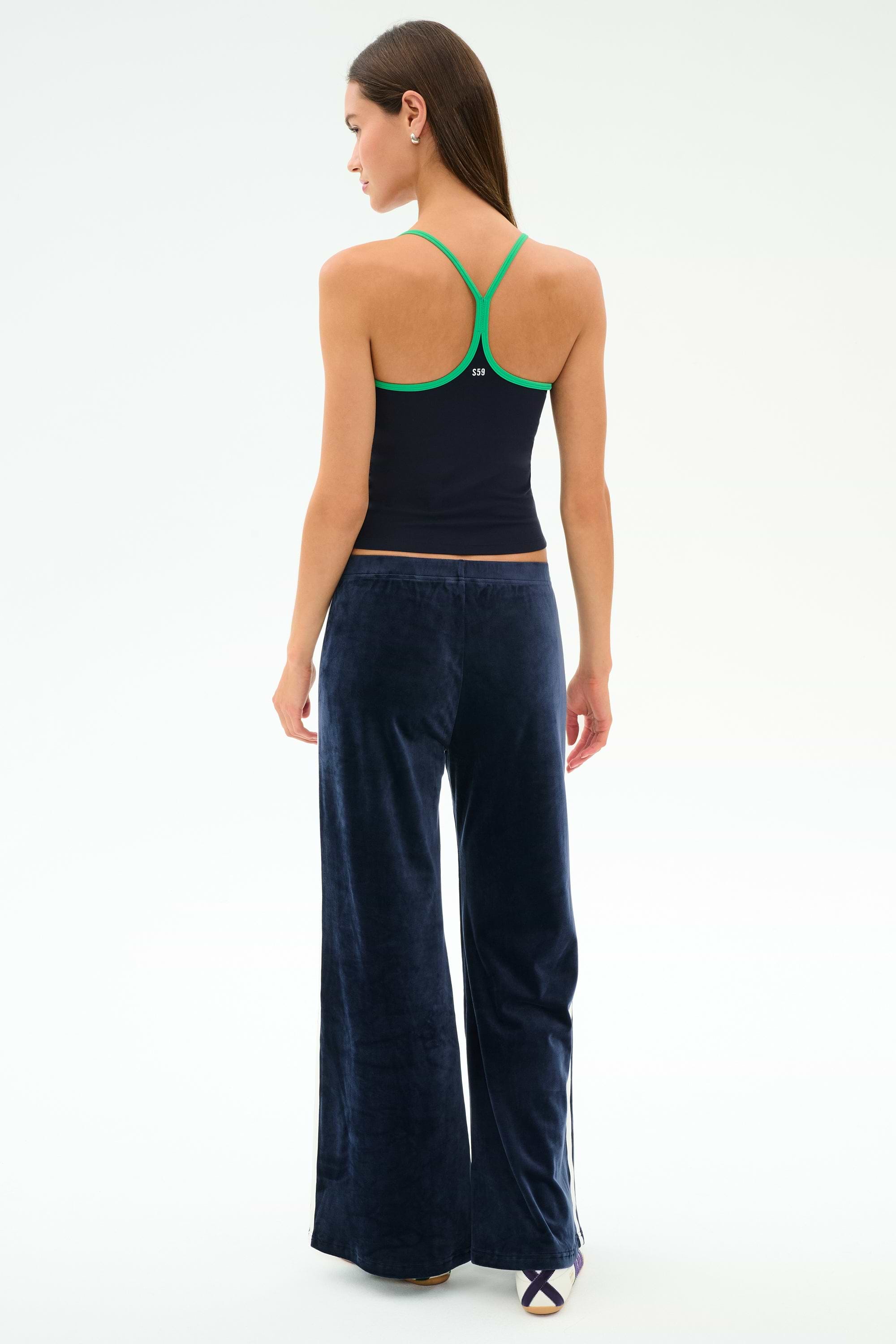 A woman stands with her back to the camera, wearing a navy racerback tank top with green trim and SPLITS59 Heritage Velour Pant - Indigo/White, featuring soft polyester-spandex fabric and a relaxed fit.