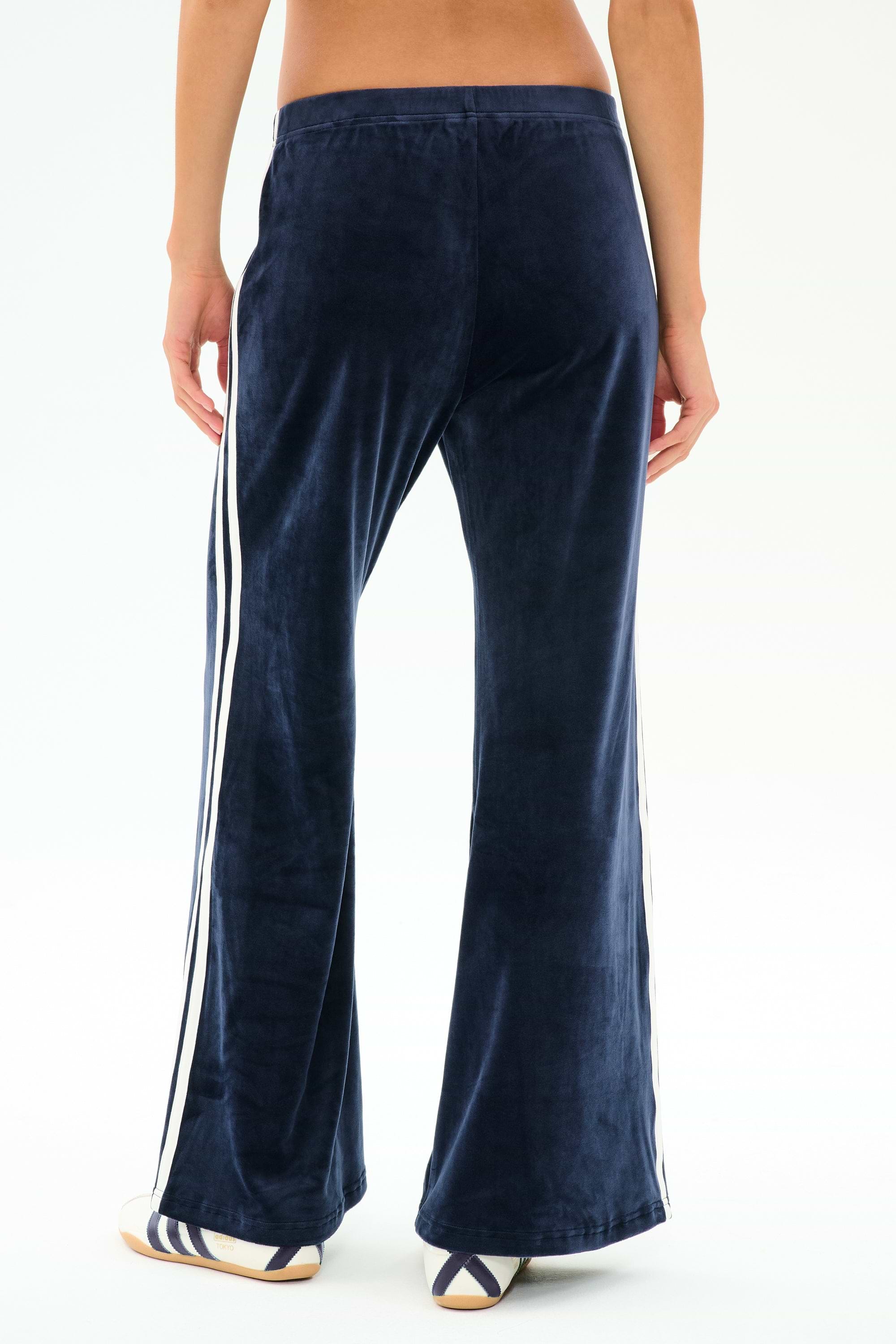 A person is shown from the back wearing SPLITS59's Heritage Velour Pant in Indigo/White, featuring white side stripes and white sneakers. The pants are made from a soft polyester blend, set against a plain white background.