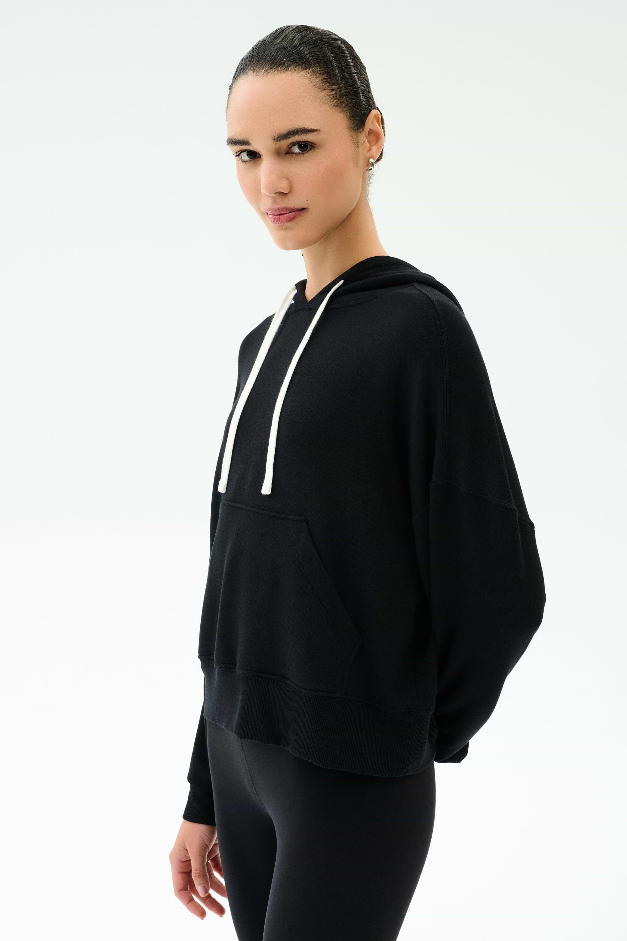 Person wearing the SPLITS59 Joey Fleece Hoodie in black—crafted from comfortable modal fabric for a soft, elevated feel—with white drawstrings and black pants, standing against a plain white background.