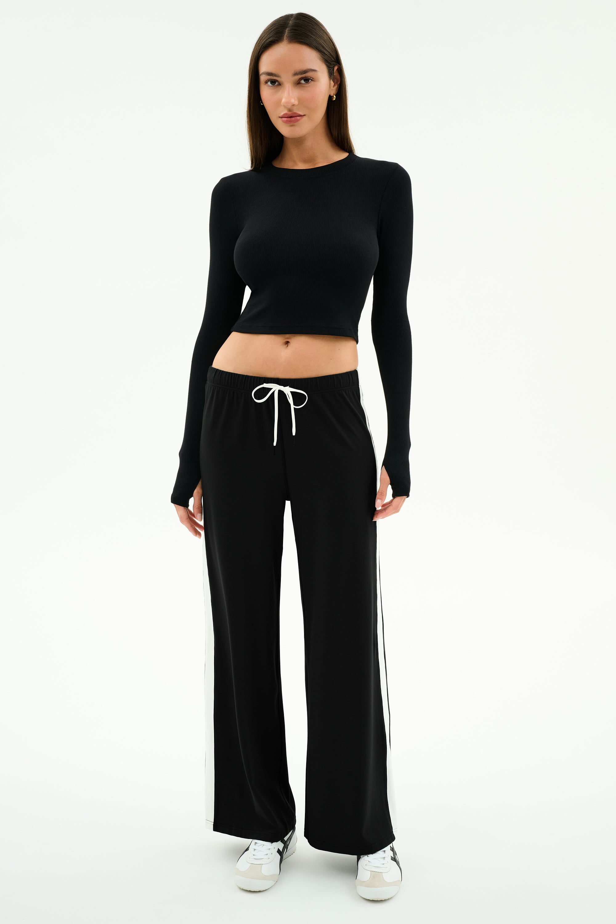A woman faces forward, wearing the SPLITS59 Louise Rib Long Sleeve Crop in black with black drawstring pants featuring white side stripes and white sneakers, set against a plain white background.