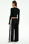 A woman stands facing away against a white background, wearing the SPLITS59 Louise Rib Long Sleeve Crop in black, paired with loose black pants with white side stripes and white sneakers.