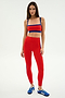 A woman wears the SPLITS59 Clare High Waist Rigor 7/8 leggings in Lotus Red/Cobalt, featuring performance fabric for workout comfort, paired with blue athletic shoes, and stands against a plain white background.