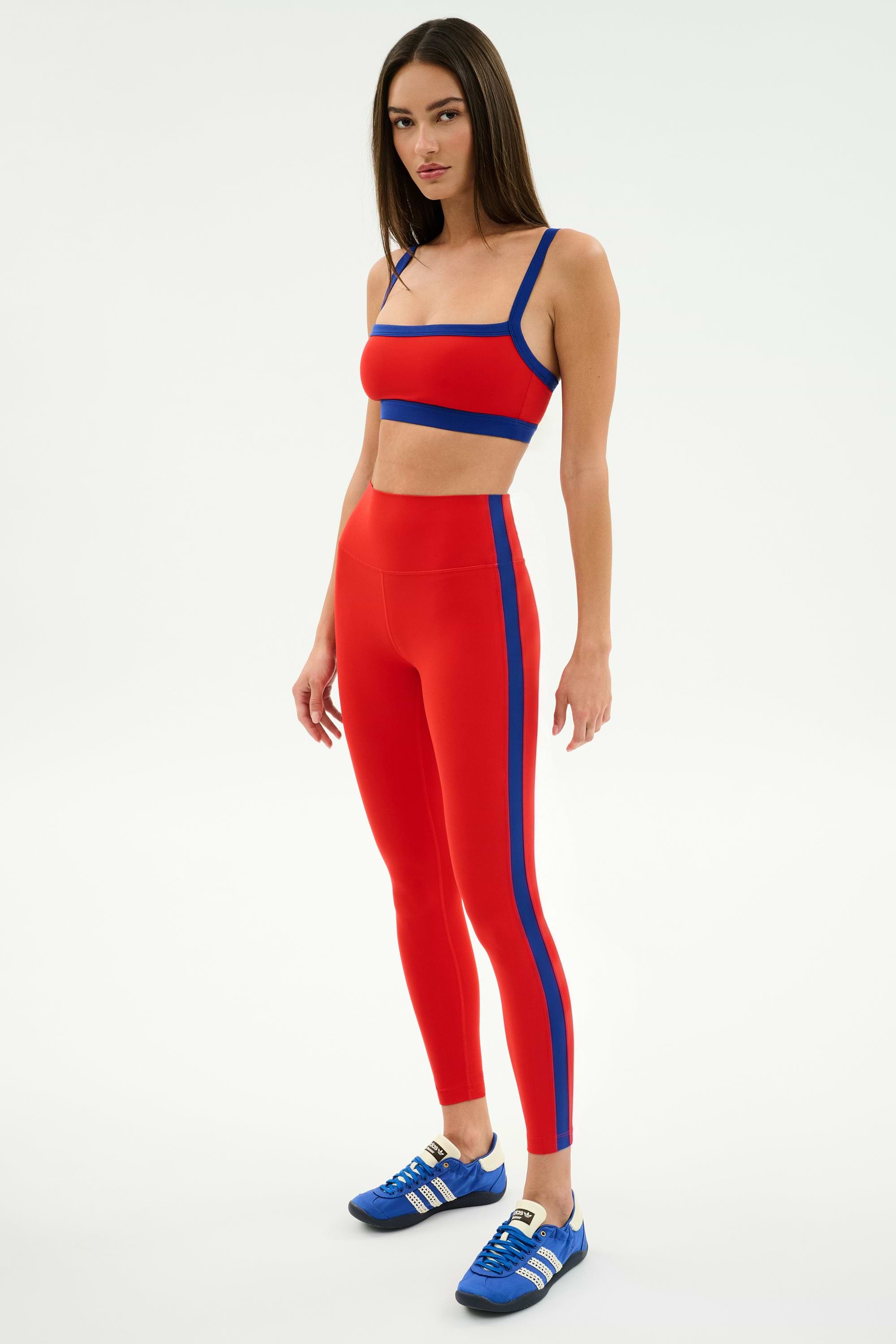 A woman wears the SPLITS59 Clare High Waist Rigor 7/8 leggings in Lotus Red/Cobalt with a matching crop top and blue sneakers, standing against a white background—ideal athletic style for your next workout.