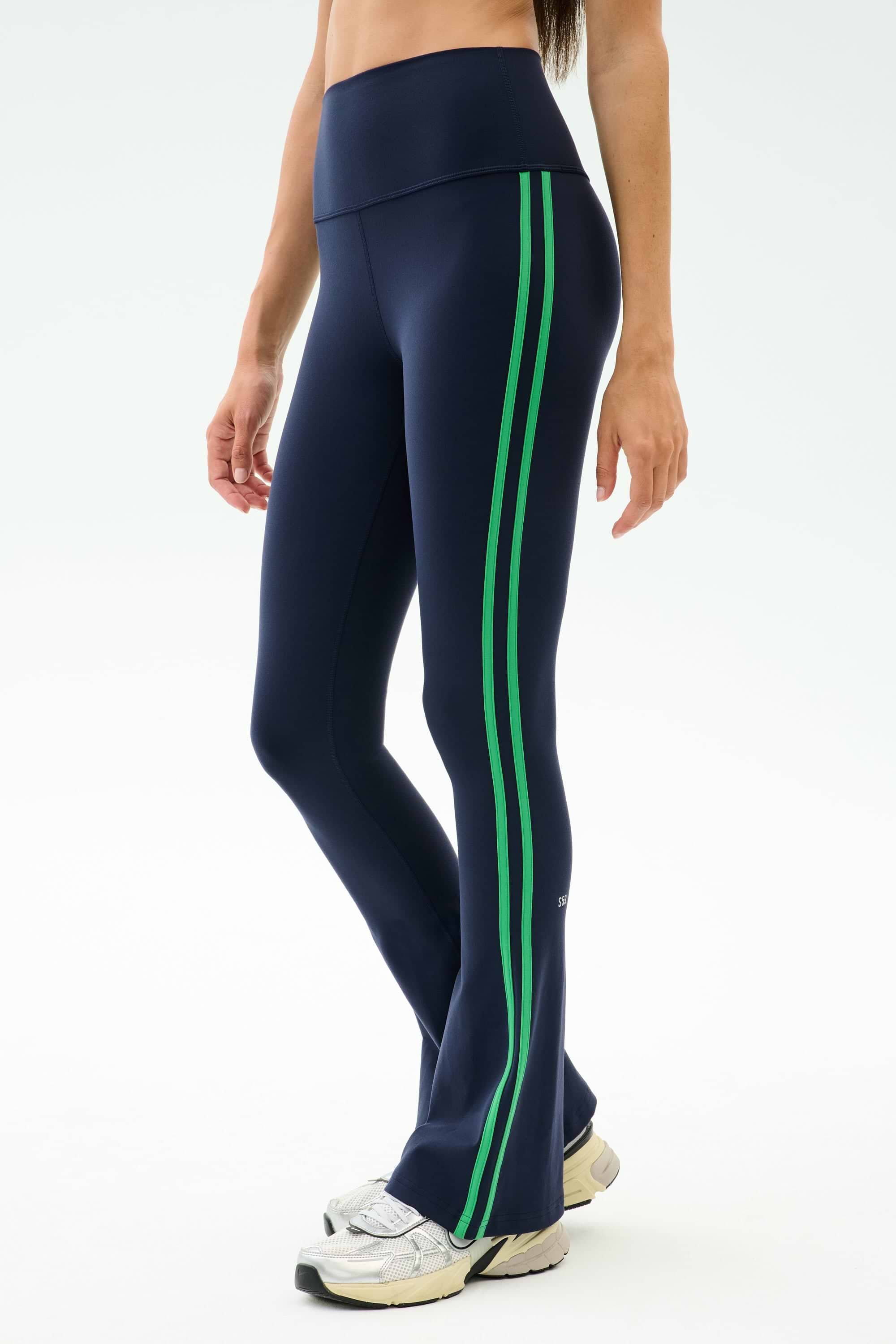 A person wears SPLITS59 Raquel High Waist Flared Legging in Indigo/Grass with white sneakers, ideal for workouts thanks to 4-way stretch fabric, standing against a plain light background.