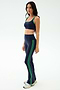 A woman stands in a studio wearing SPLITS59’s Raquel High Waist Flared Legging in Indigo/Grass, featuring green stripes and 4-way stretch fabric for ultimate comfort—perfect for any workout. She pairs them with a matching sports bra and white sneakers.